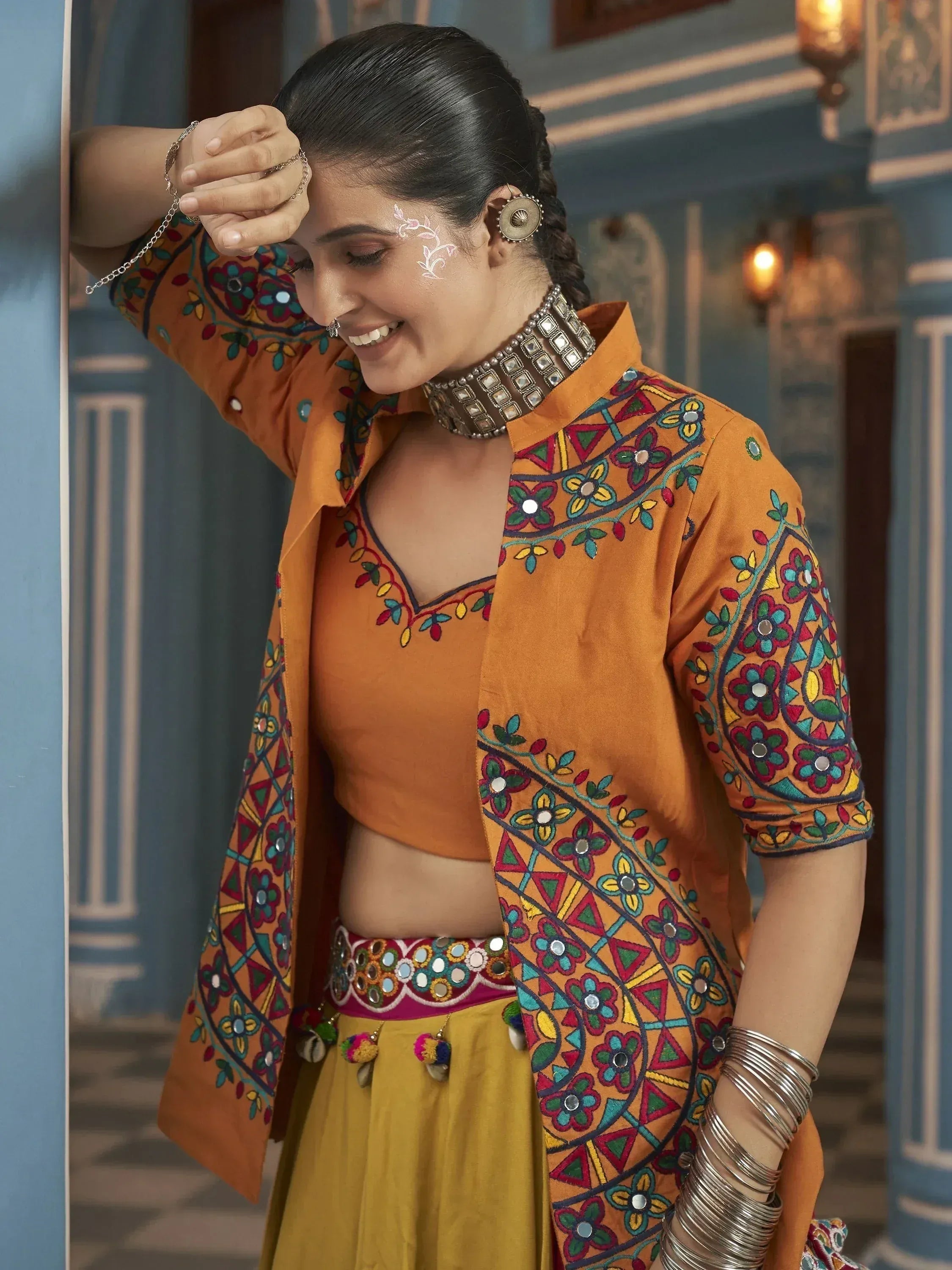 Woman in vibrant orange embroidered Indian ethnic wear with mirror work, paired with yellow skirt and traditional jewelry