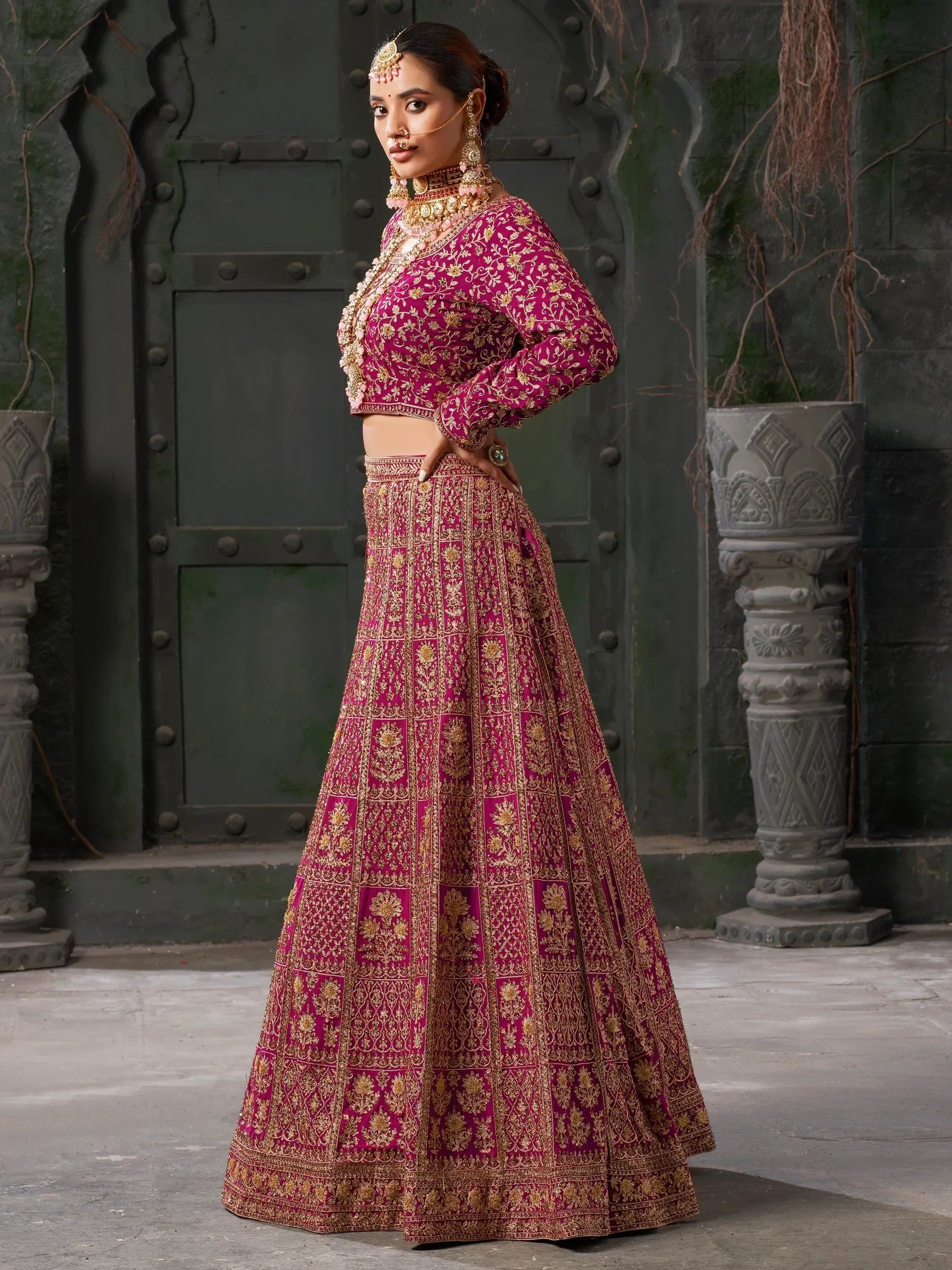 Woman in magenta and gold embroidered lehenga choli with traditional jewelry, perfect for Indian weddings or festive occasions.