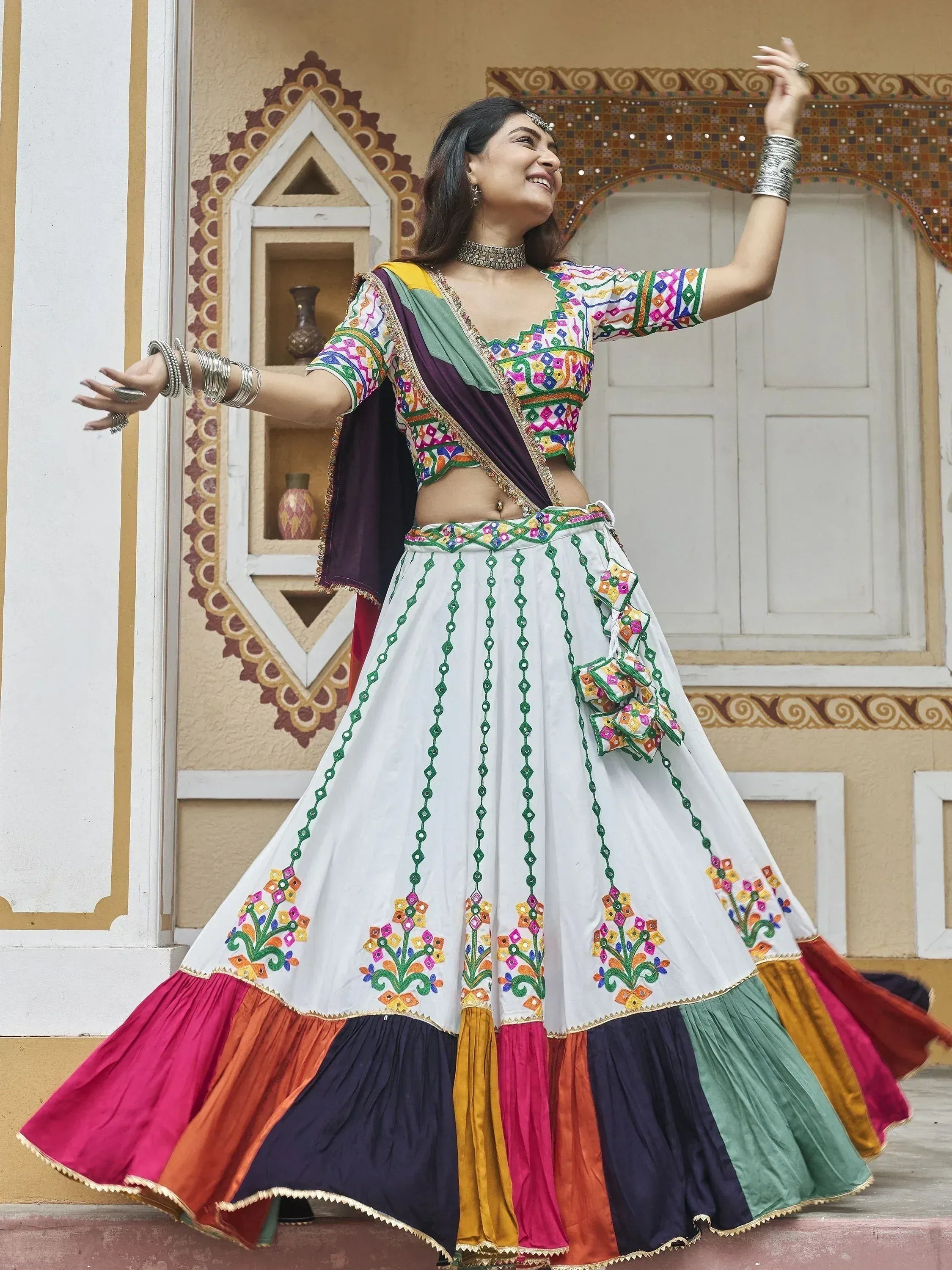 Woman in colorful embroidered lehenga choli with mirror work, dancing joyfully against ornate Indian architecture