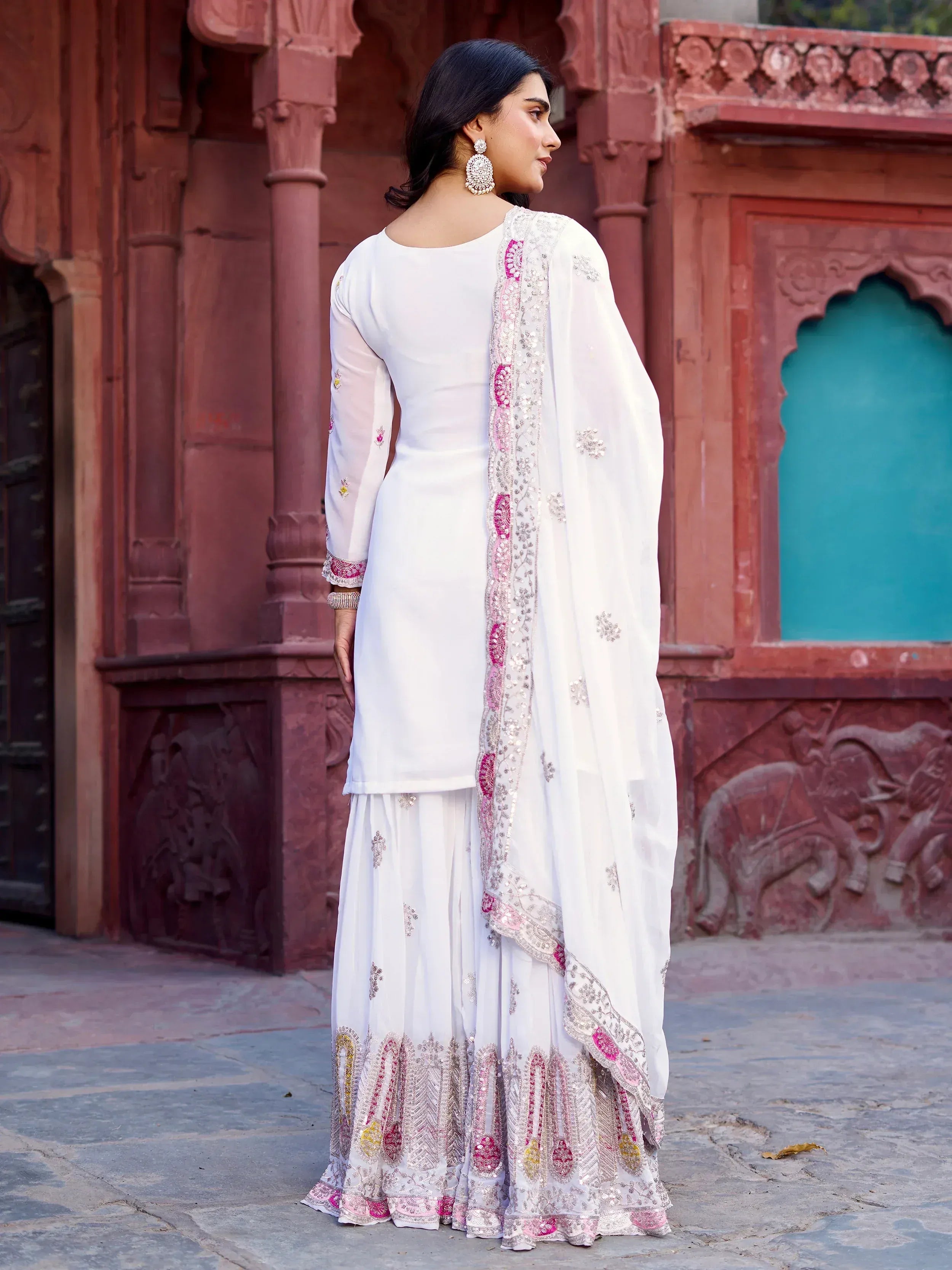 White embroidered salwar kameez with pink and gold detailing, styled with dupatta against red stone architecture