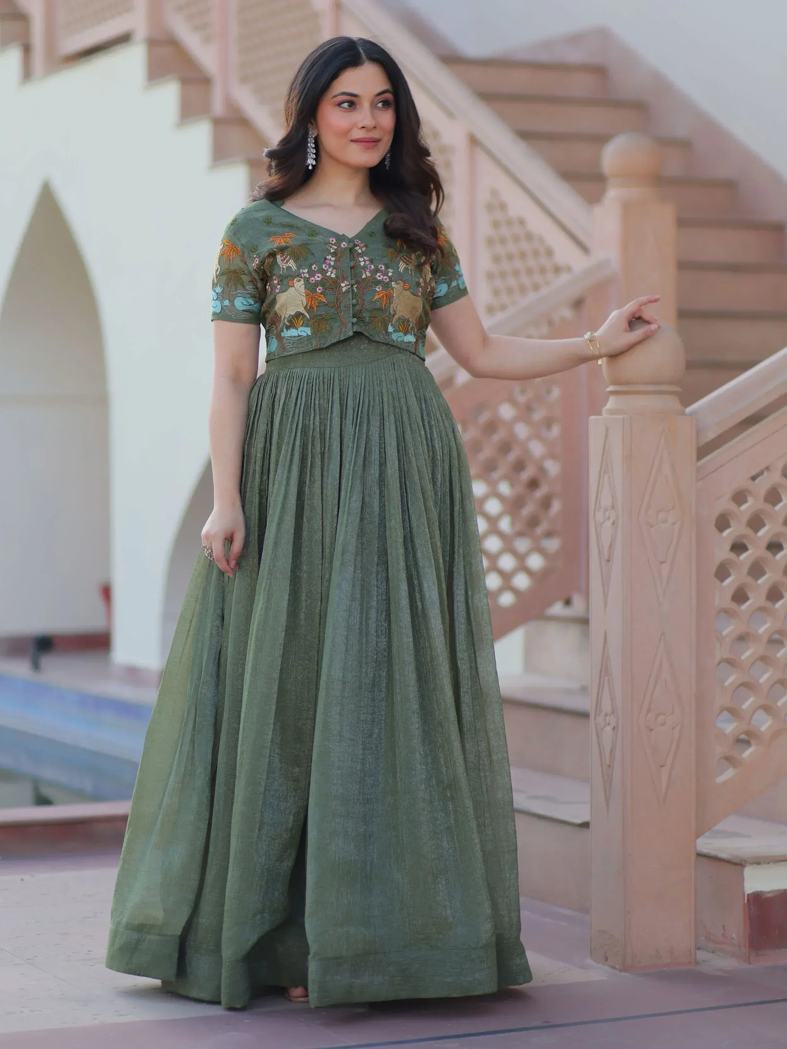 Woman in olive green embroidered ethnic gown with floral and animal motifs, standing by staircase
