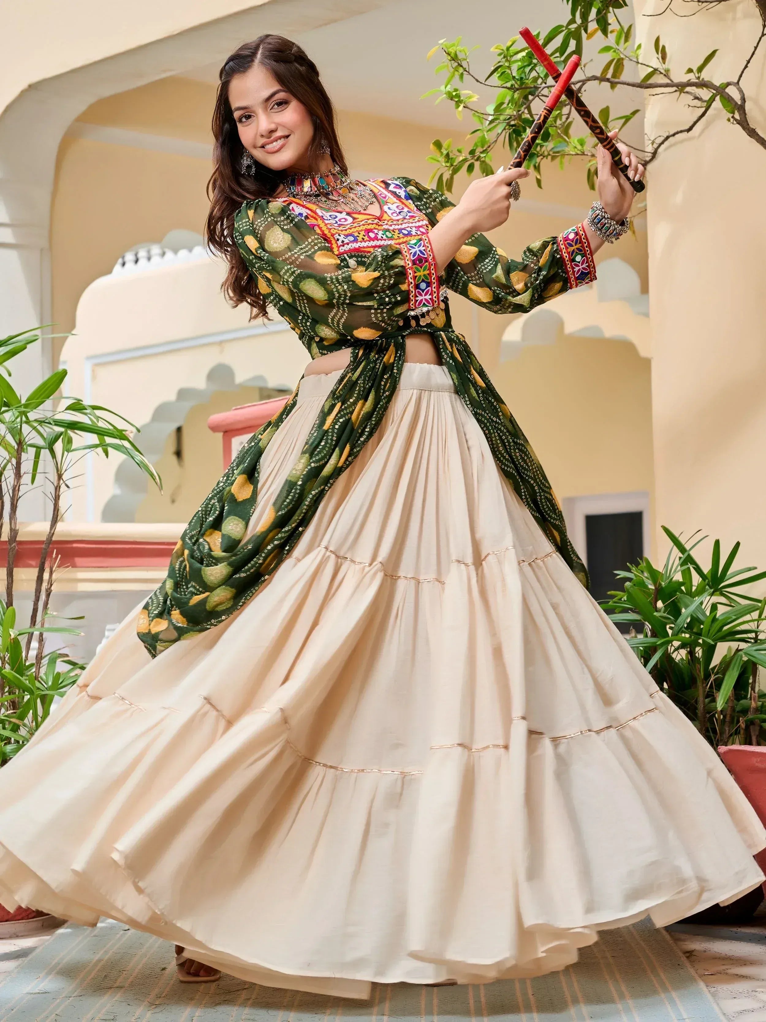 Woman in vibrant green embroidered top and cream pleated skirt, holding dandiya sticks for festive Indian wear