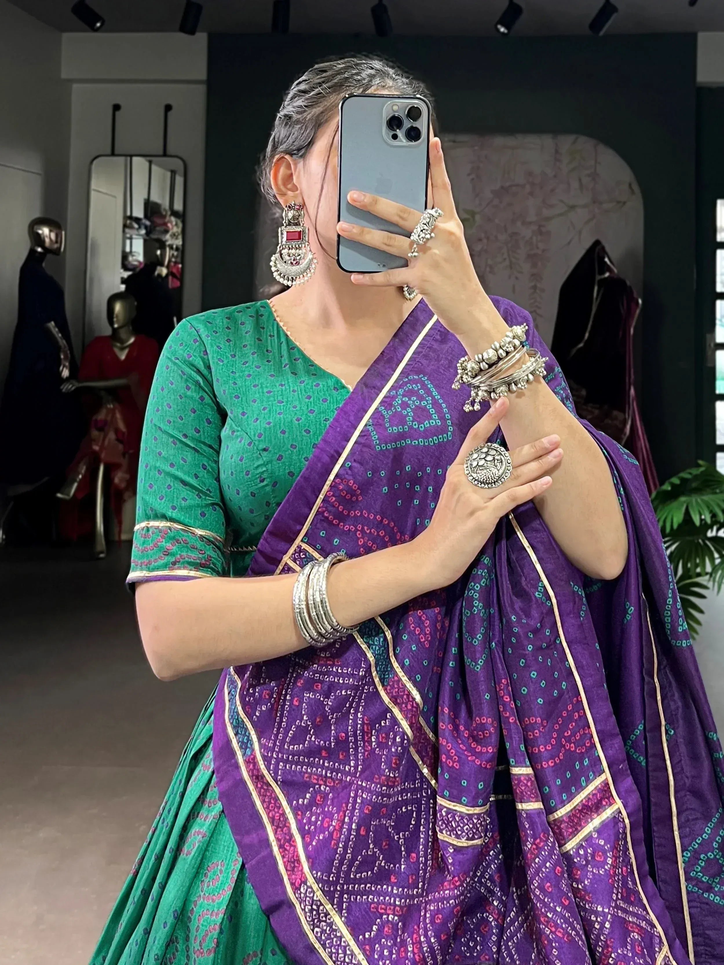 Woman in purple and green printed saree with ethnic jewelry, taking mirror selfie in boutique