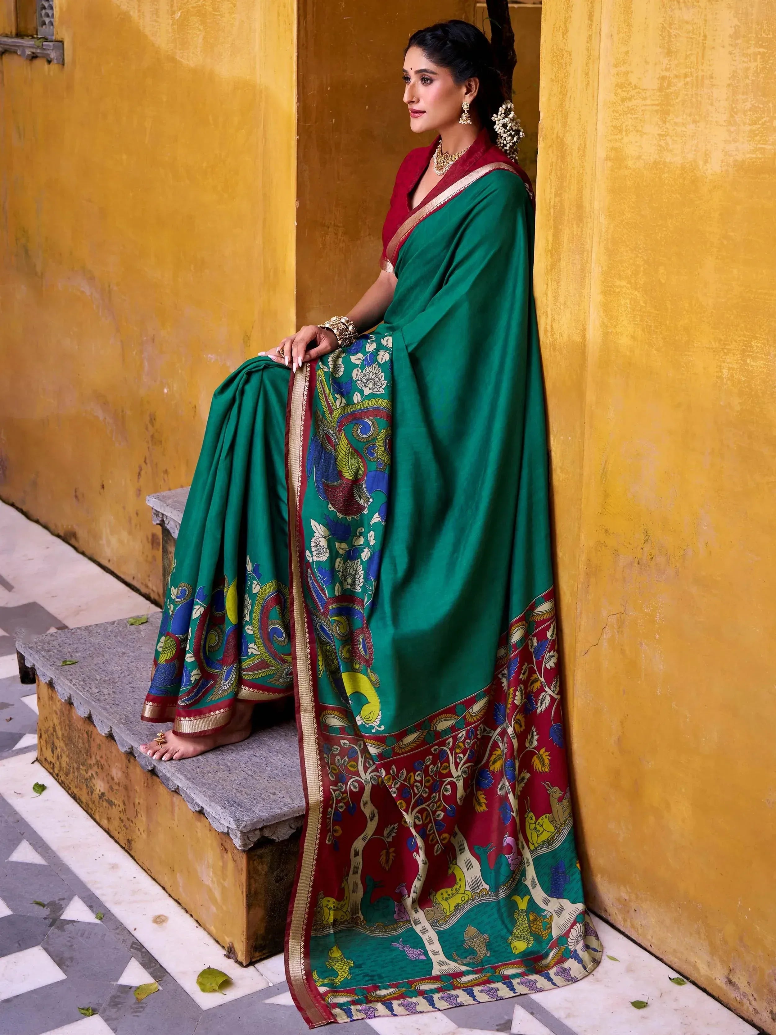 Woman in green and red printed saree with peacock design, ethnic Indian wear against yellow wall