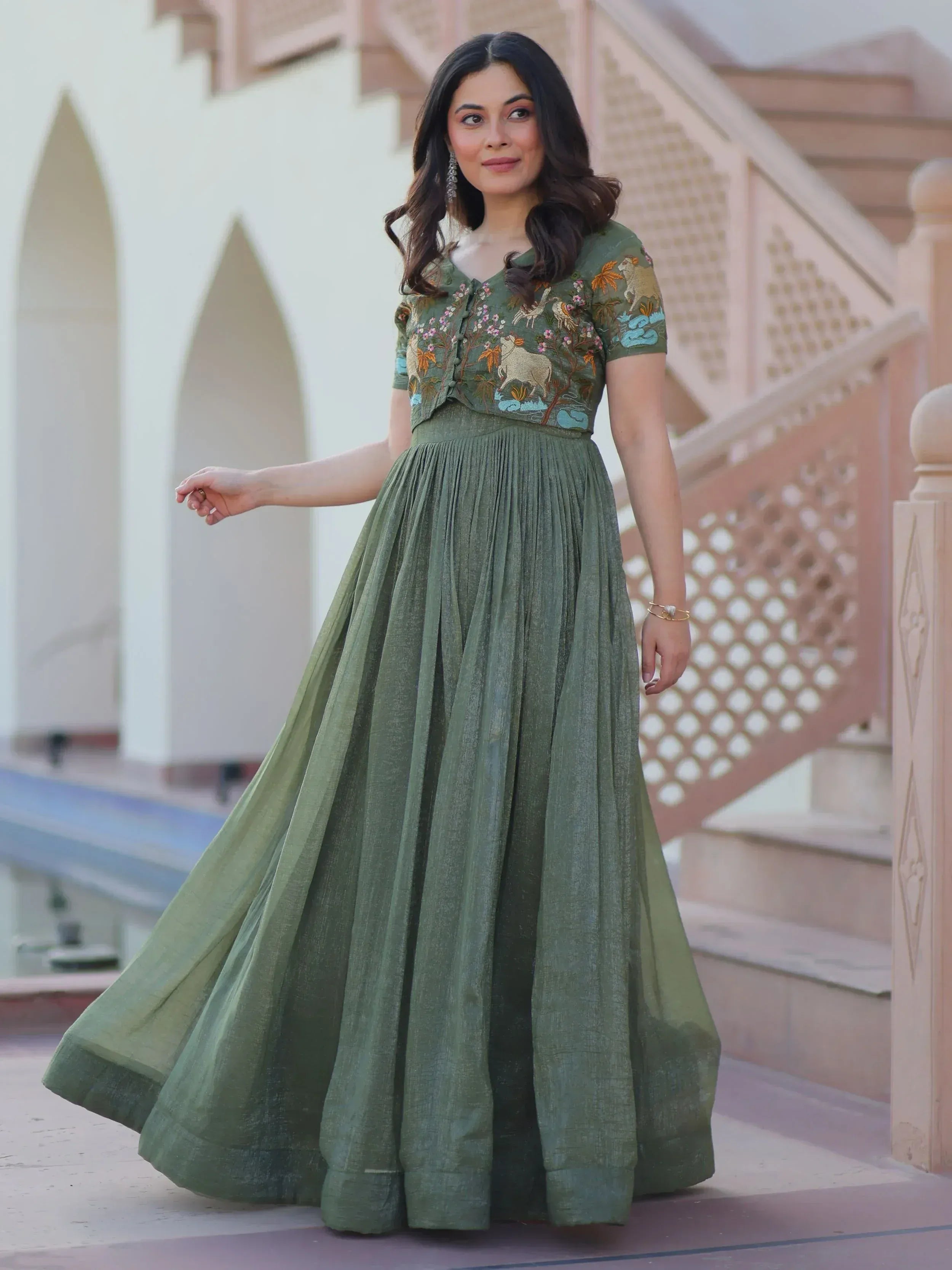 Woman in olive green embroidered ethnic gown with animal motifs, pleated skirt, standing near arched architecture