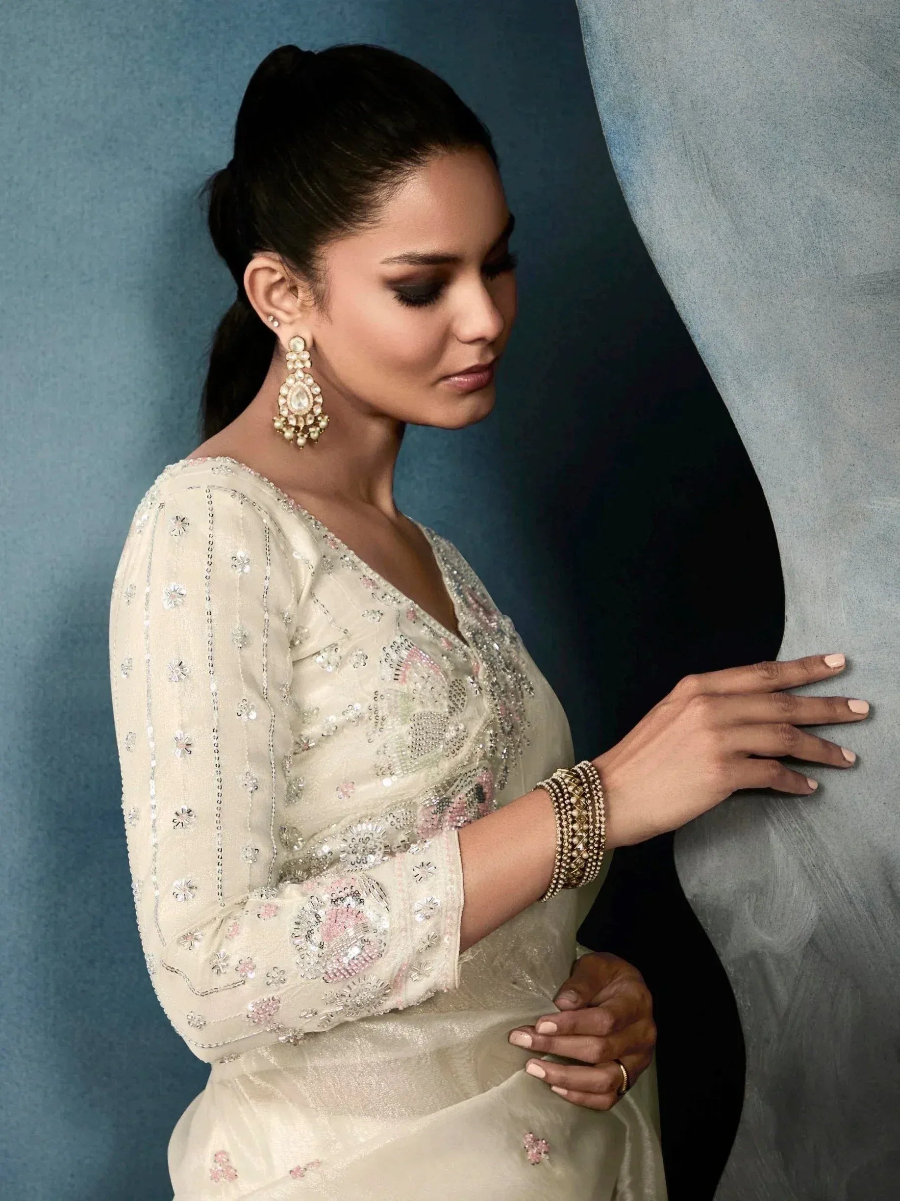 Elegant woman in cream embroidered ethnic gown with floral beadwork, statement earrings, and bangles against blue backdrop