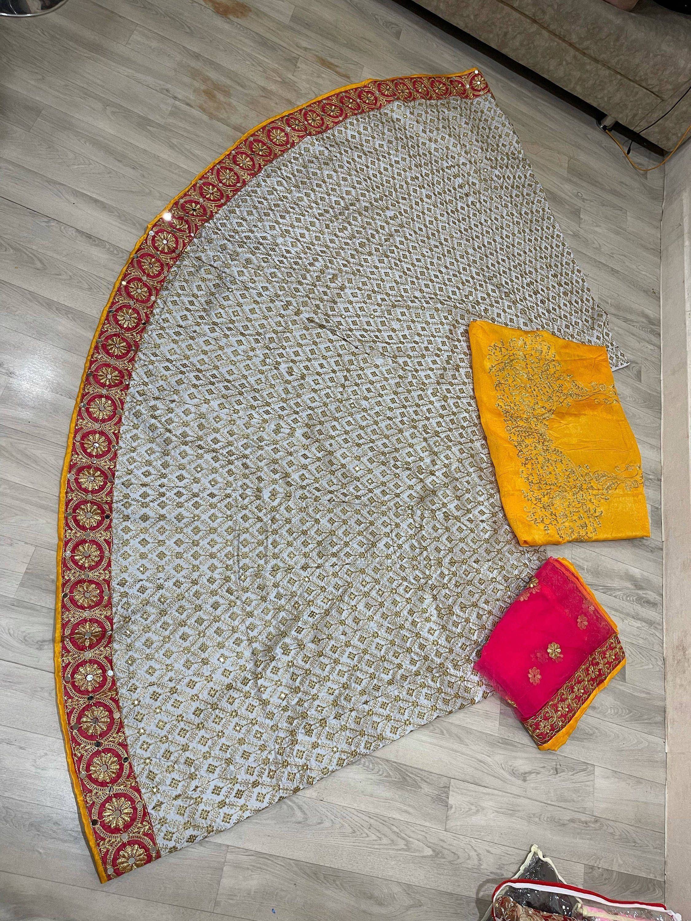 White and gold embroidered lehenga with red border, yellow blouse, and pink dupatta, laid on wooden floor