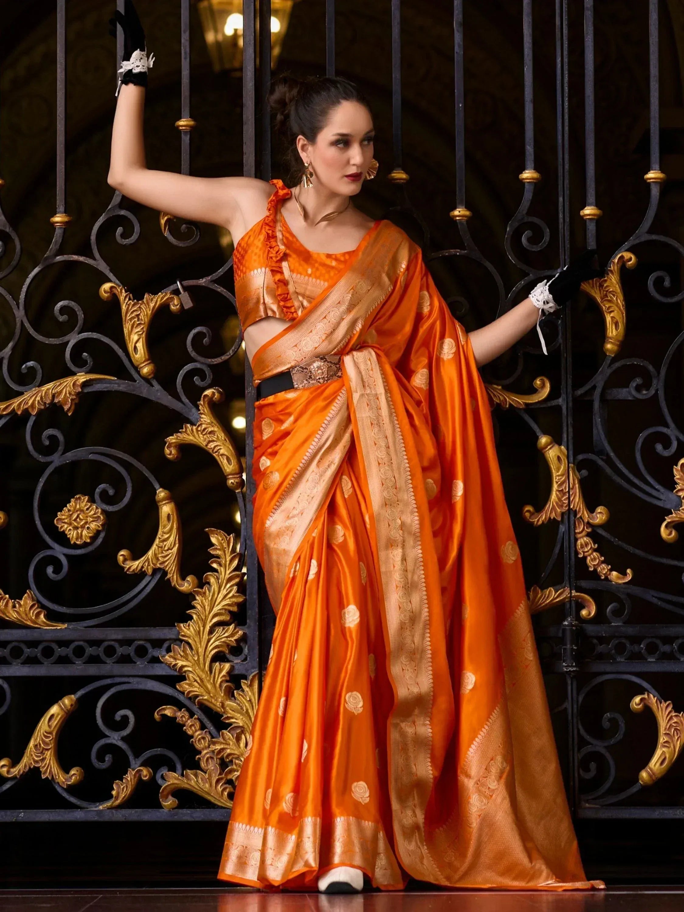 Orange silk saree with gold embroidery, paired with ruffled blouse, worn by woman near ornate gate