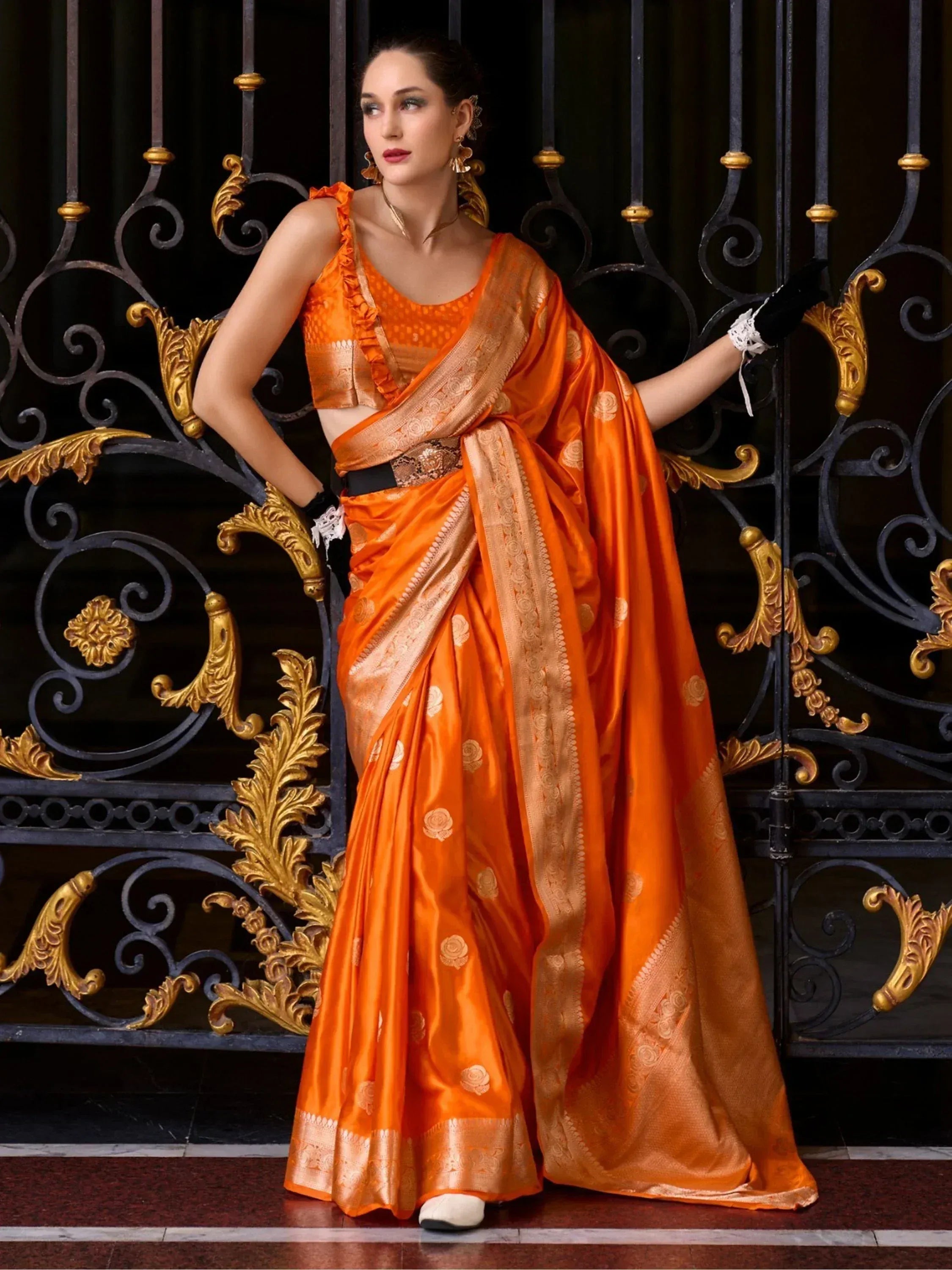 Orange designer saree with gold motifs, ruffled blouse, black gloves, against ornate gate