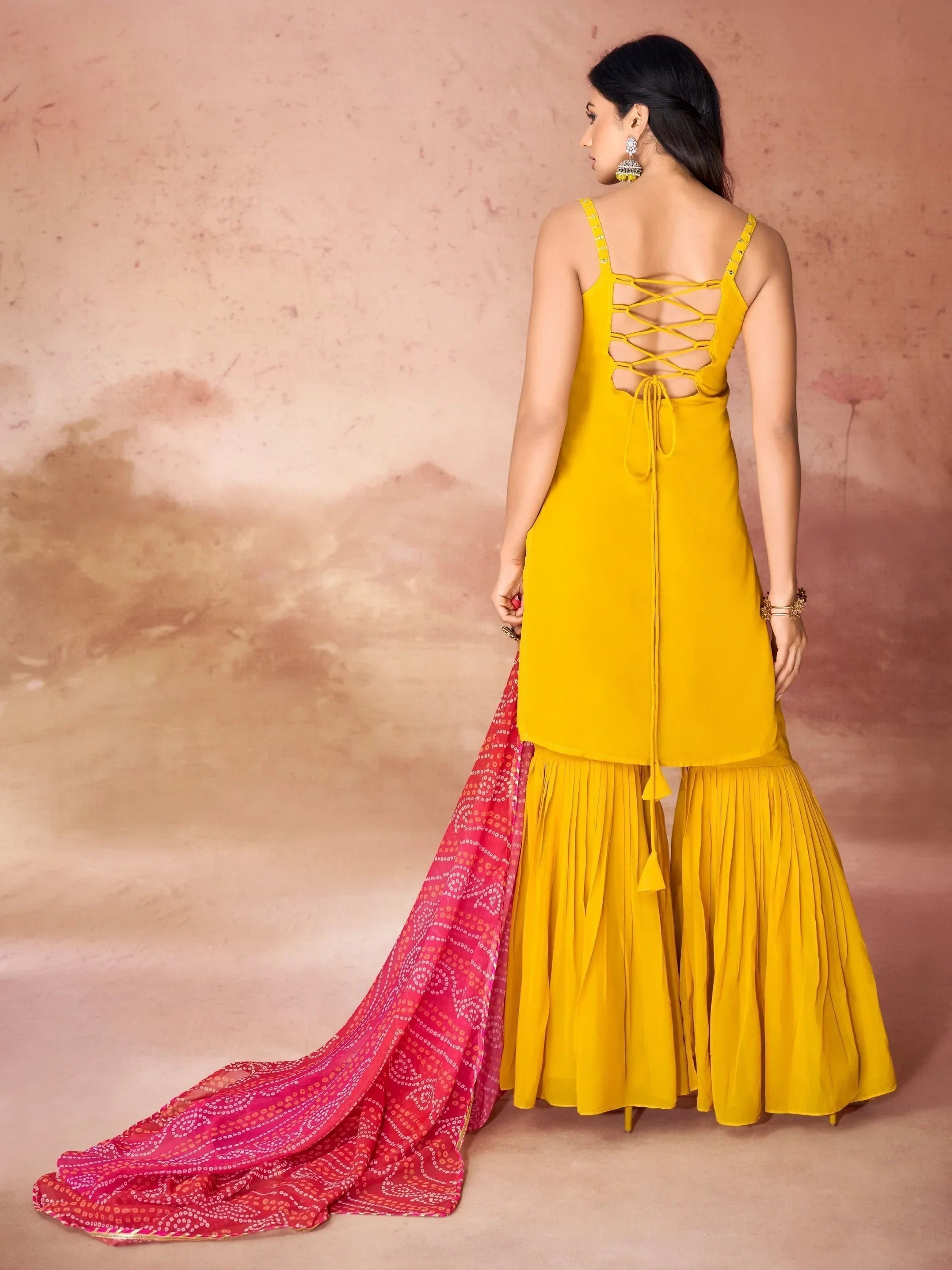 Woman in yellow sharara suit with back lace-up design and pink bandhani dupatta