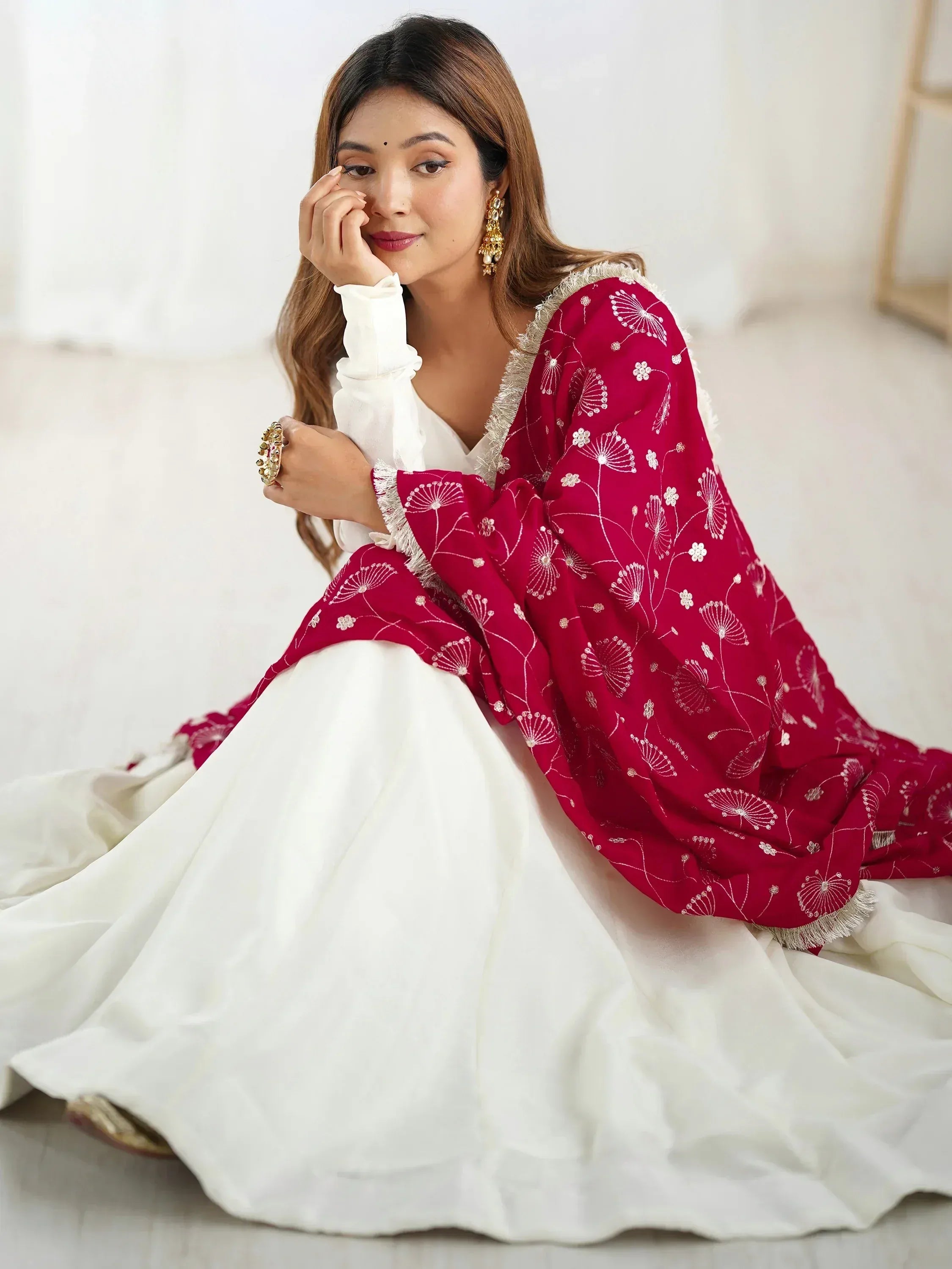Woman in white ethnic gown with red floral embroidered dupatta, gold jewelry, sitting indoors