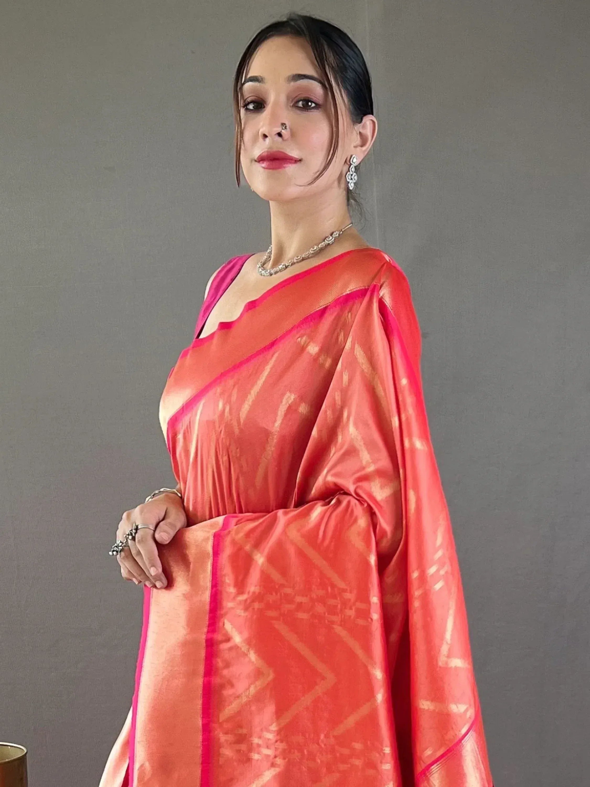 Woman in vibrant coral saree with gold geometric patterns and pink border, styled with ethnic jewelry
