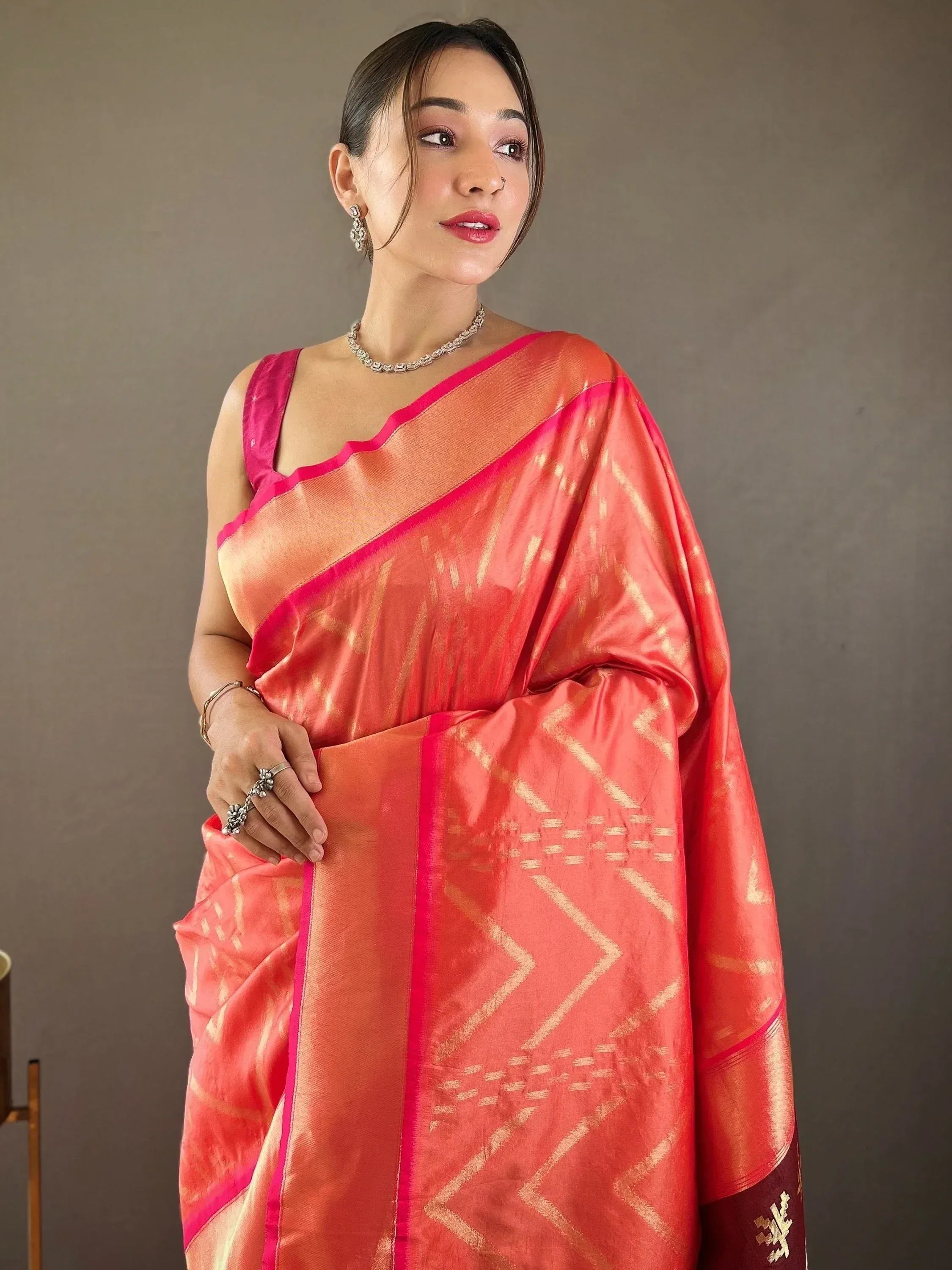Woman in coral pink saree with gold geometric patterns and vibrant pink border, styled with ethnic jewelry