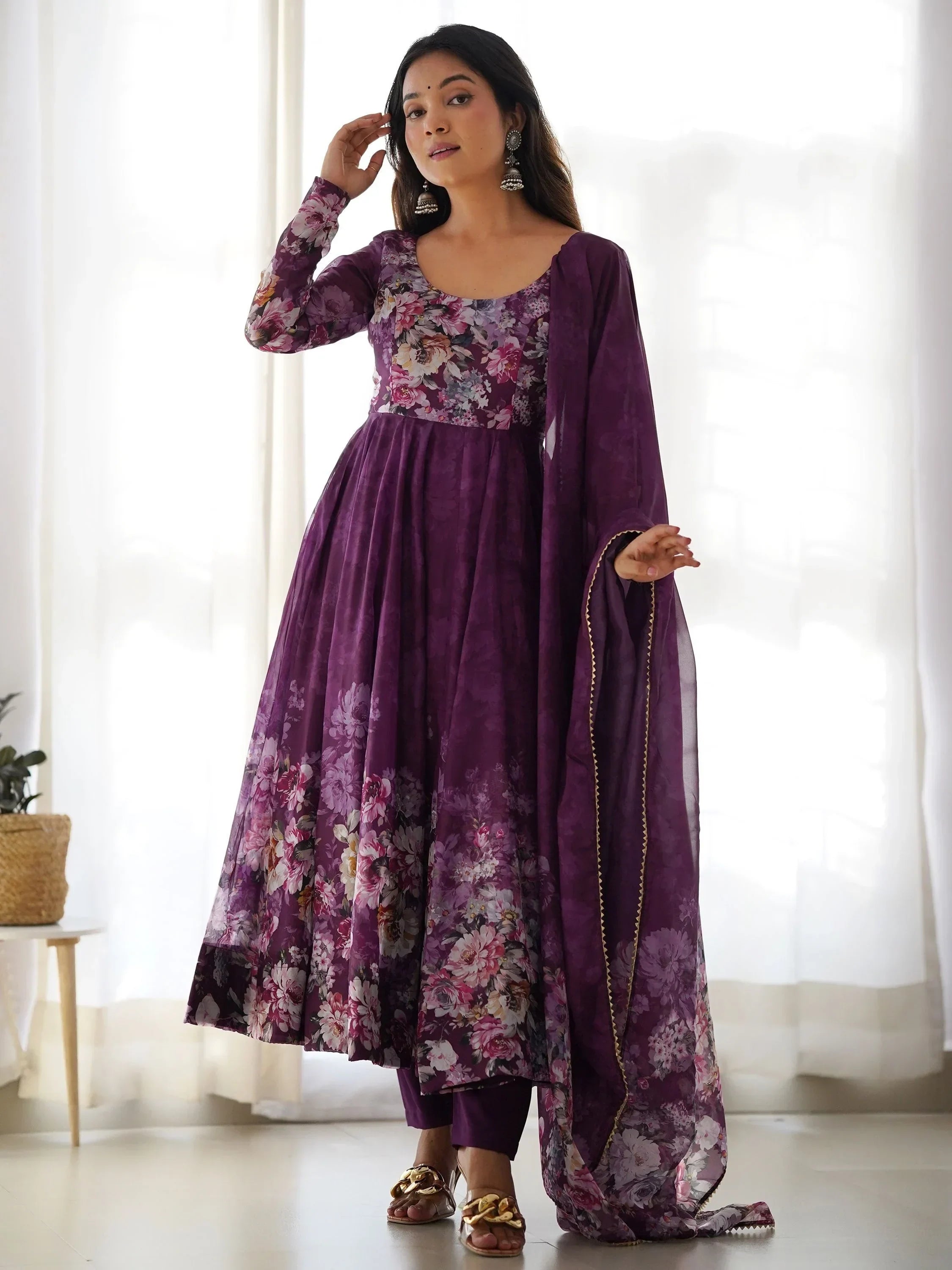 Woman wearing a purple floral Anarkali suit with dupatta and gold sandals in bright room