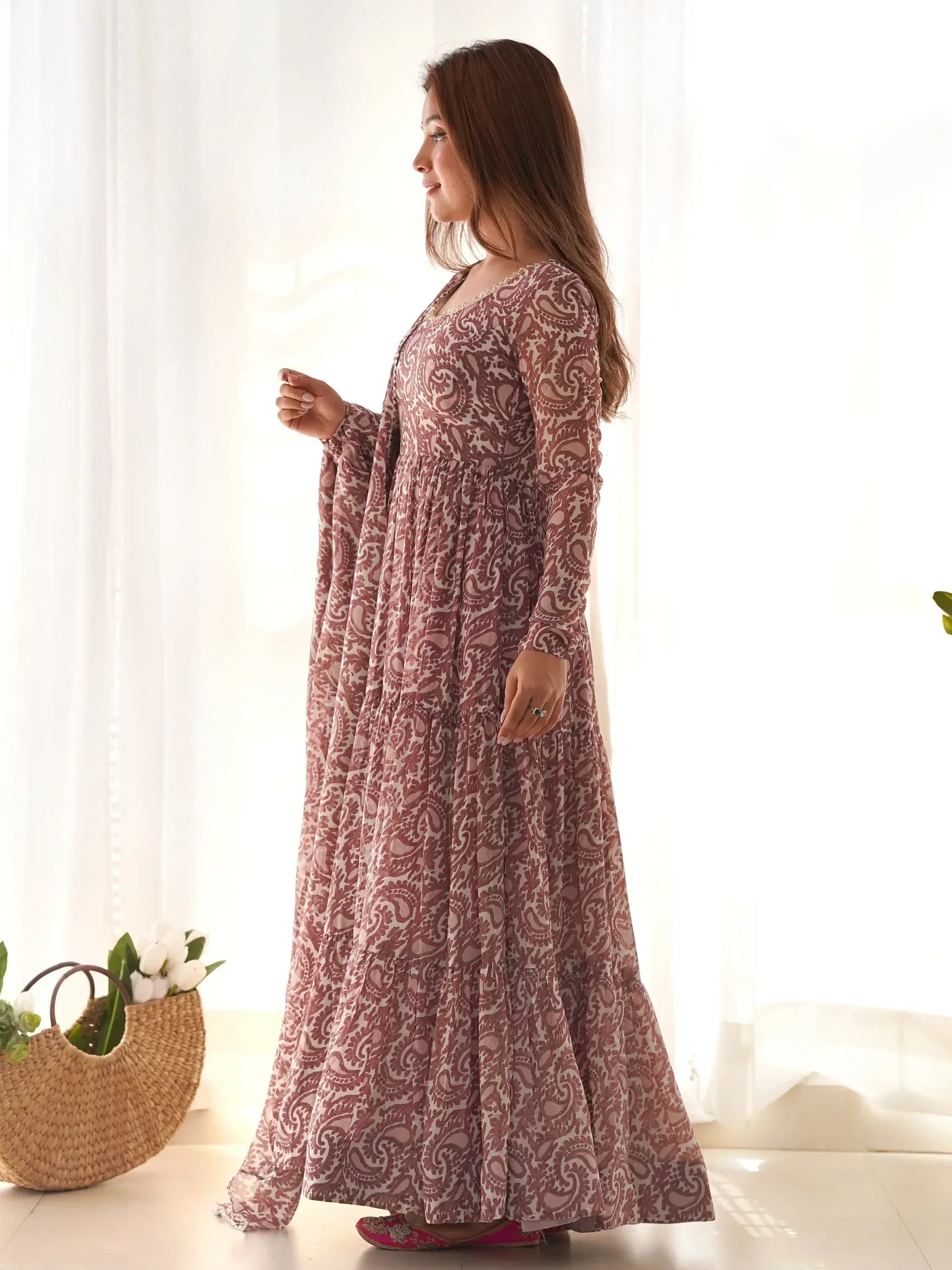 Woman in a mauve printed ethnic gown standing indoors by white curtains