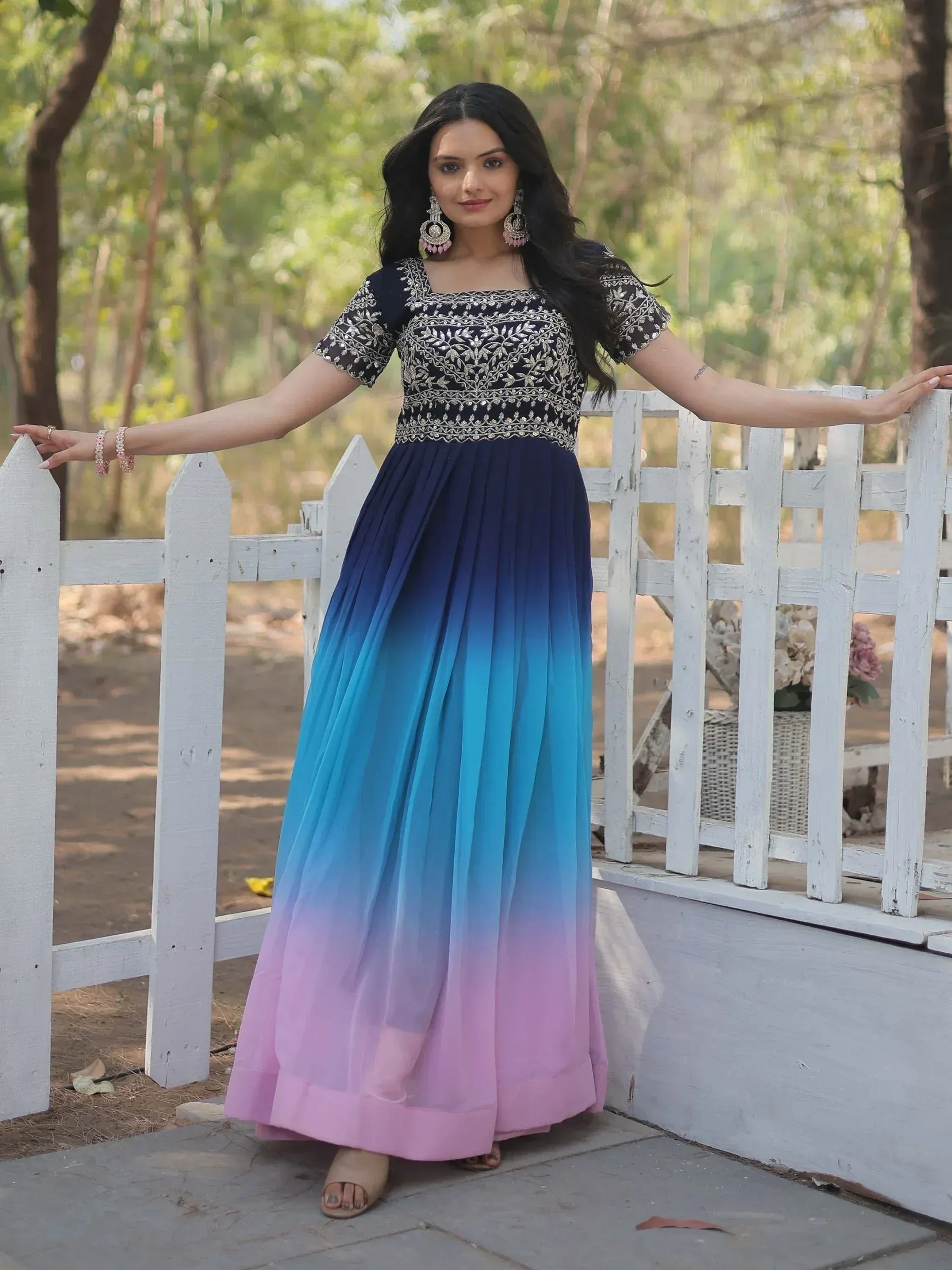 Woman in embroidered black top with ombre blue to pink pleated ethnic gown, standing by white fence