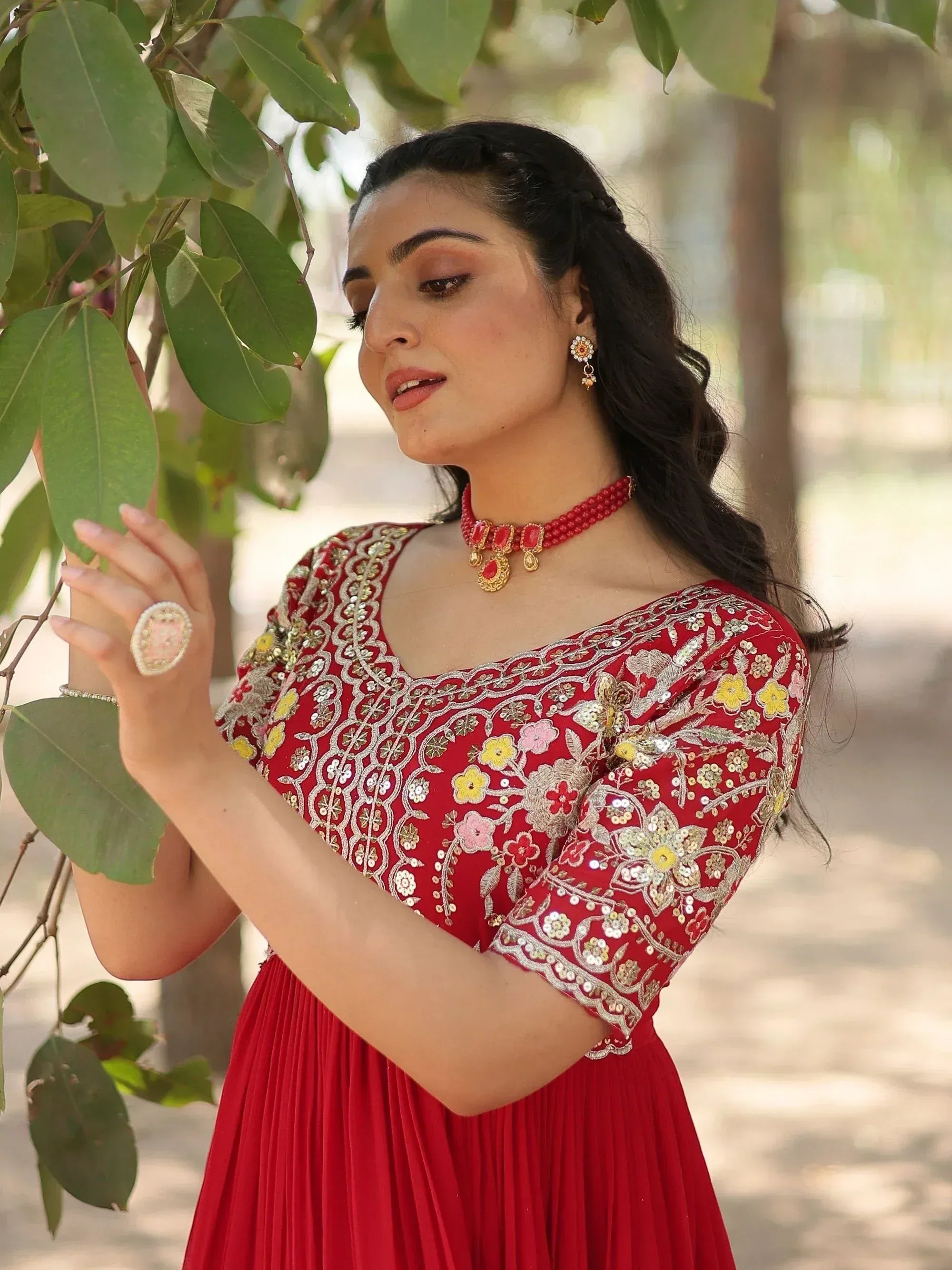 Woman in red embroidered ethnic gown with floral design, wearing matching jewelry under green leaves
