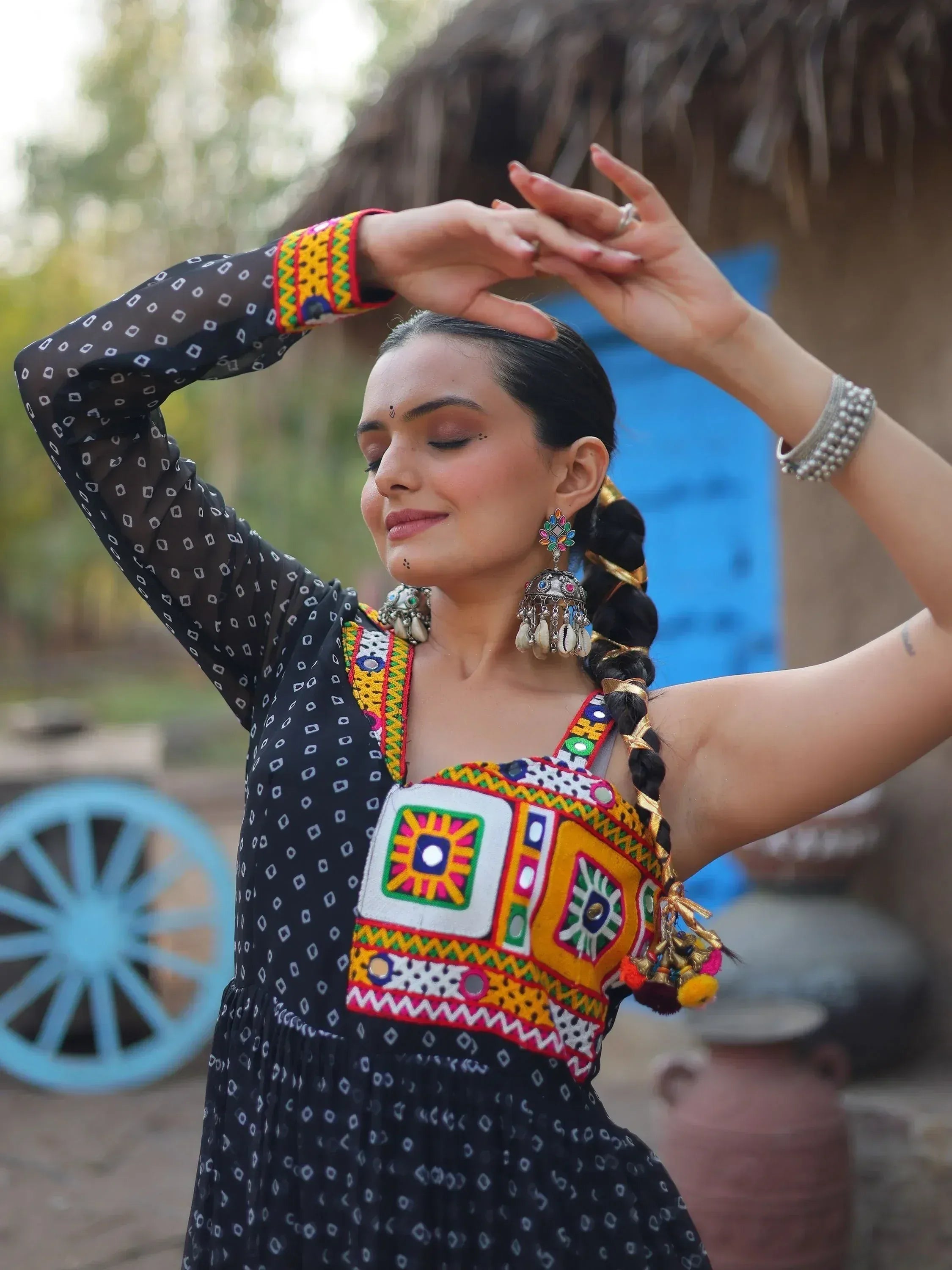 Woman in black ethnic dress with colorful mirror work, traditional jewelry, and braided hair, posing outdoors.