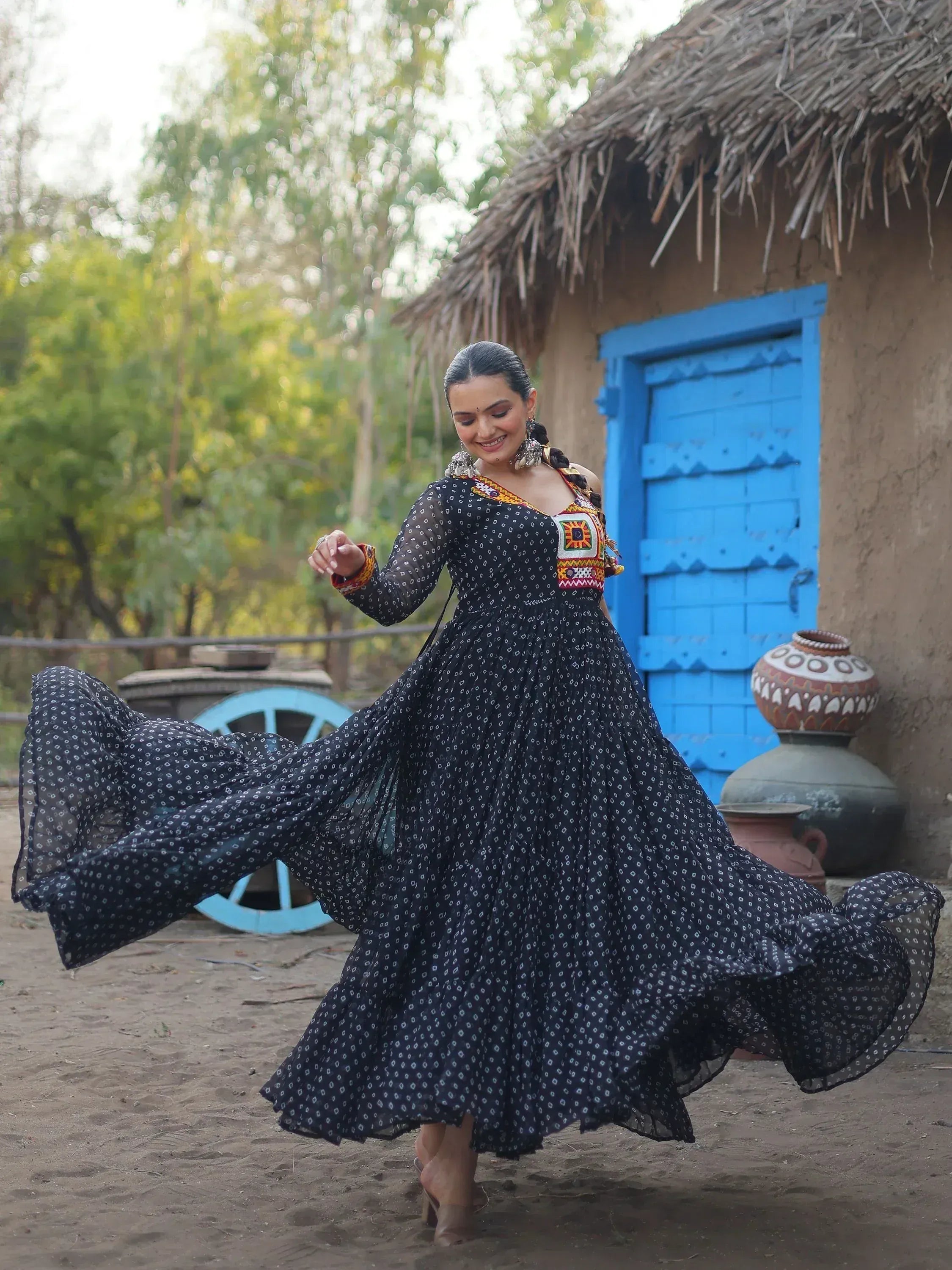 Woman twirling in black printed ethnic gown with mirror work, village backdrop