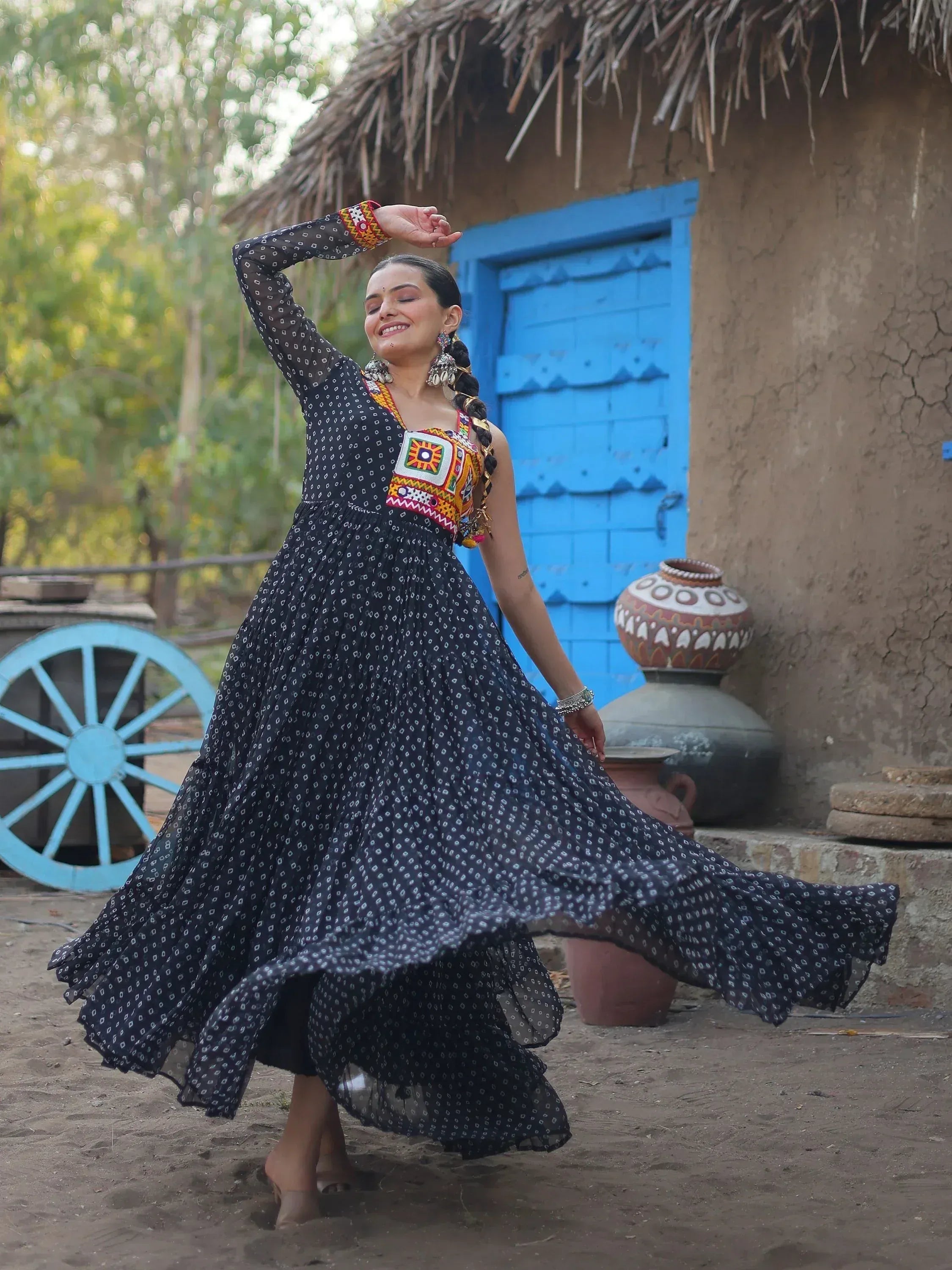 Woman in black printed ethnic gown with colorful embroidery, dancing near rustic hut with blue door.