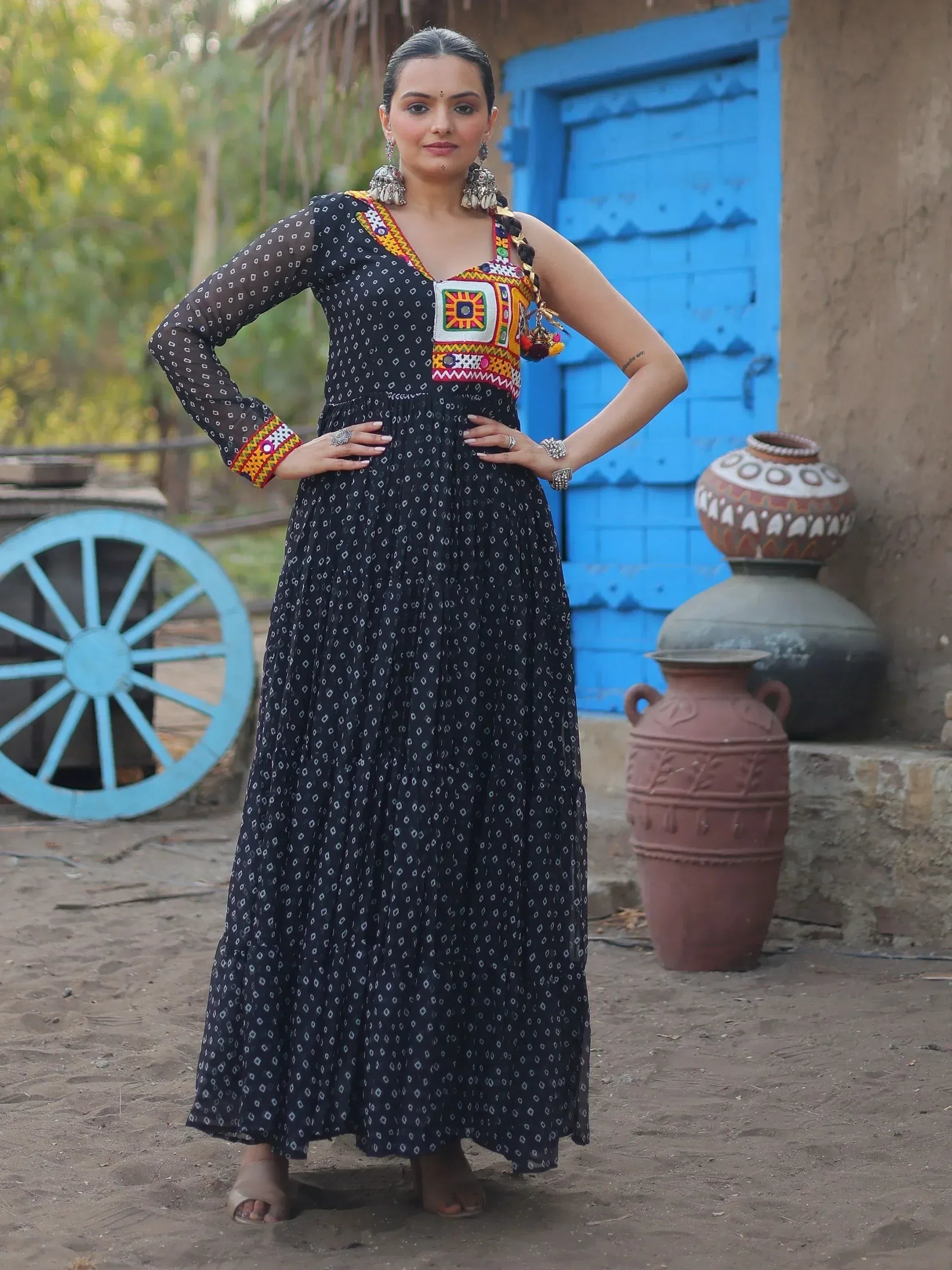 Woman in black ethnic gown with colorful embroidery, standing near blue door and clay pots