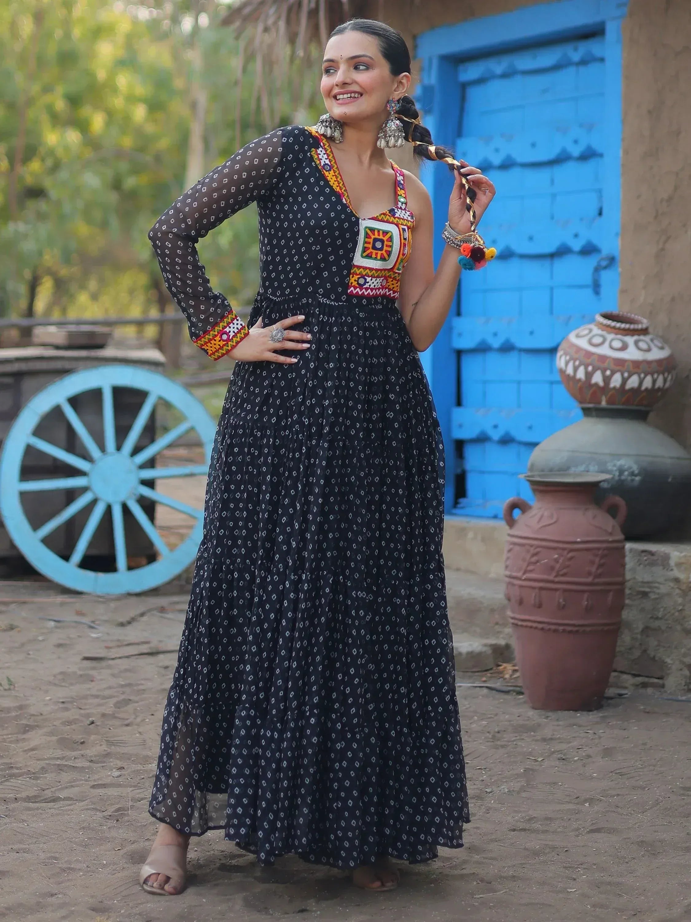 Woman in black printed ethnic gown with colorful embroidery, standing near blue door and clay pots