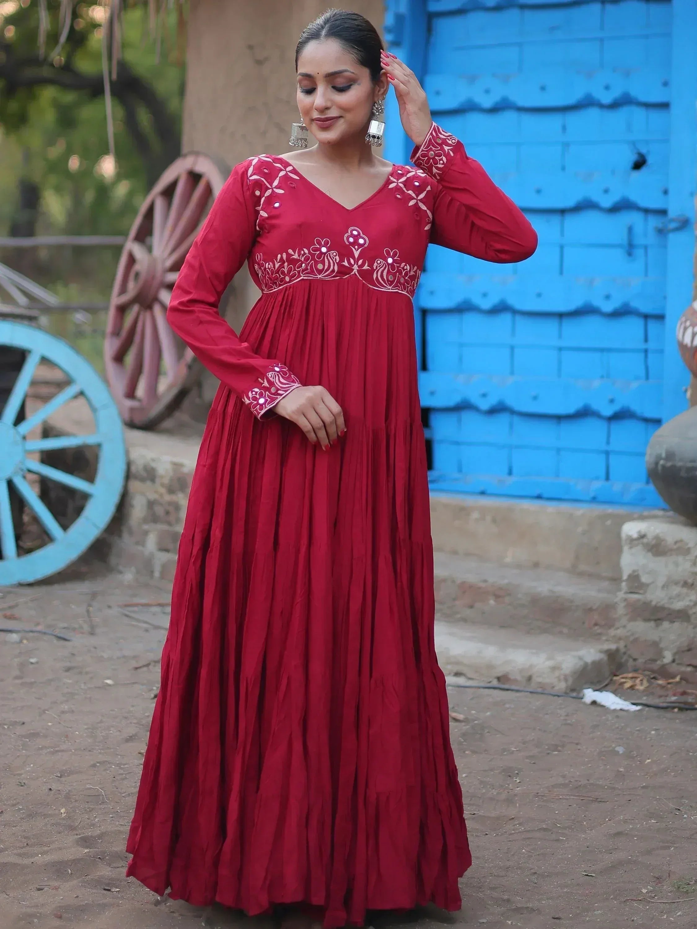 Red ethnic gown with white embroidery, long sleeves, and pleated skirt, styled with jhumka earrings against blue door backdrop