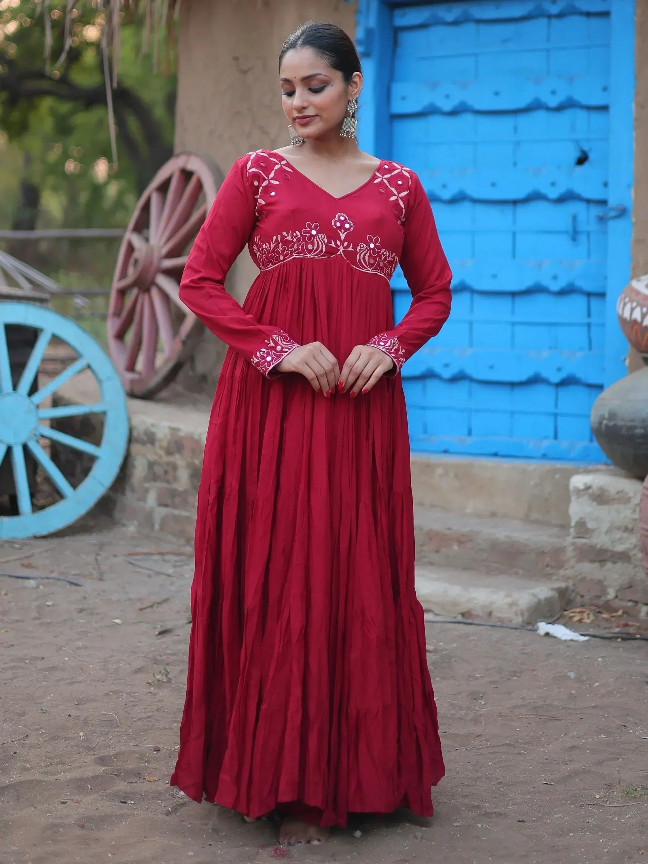 Red ethnic gown with white embroidery, long sleeves, and pleated skirt, styled with jhumka earrings against rustic blue door backdrop