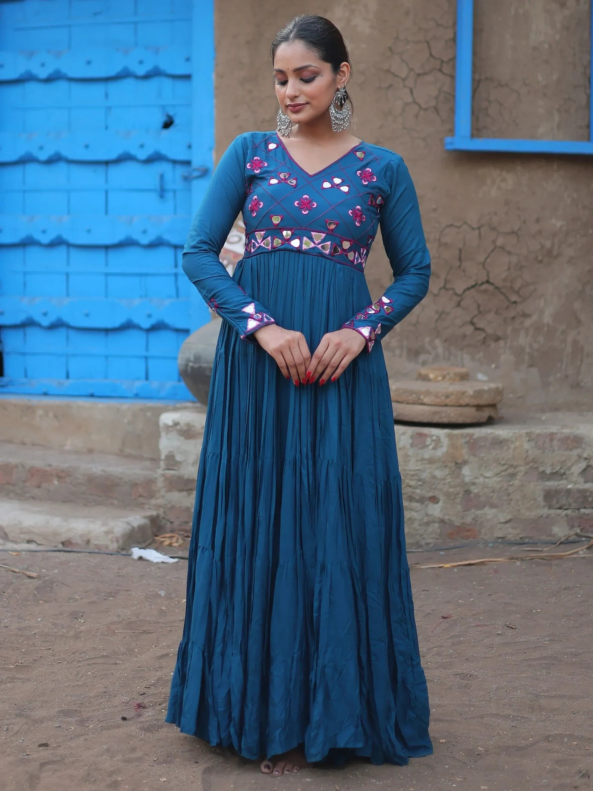 Woman wearing a blue ethnic gown with mirror work, long sleeves, and floral embroidery, standing outdoors.