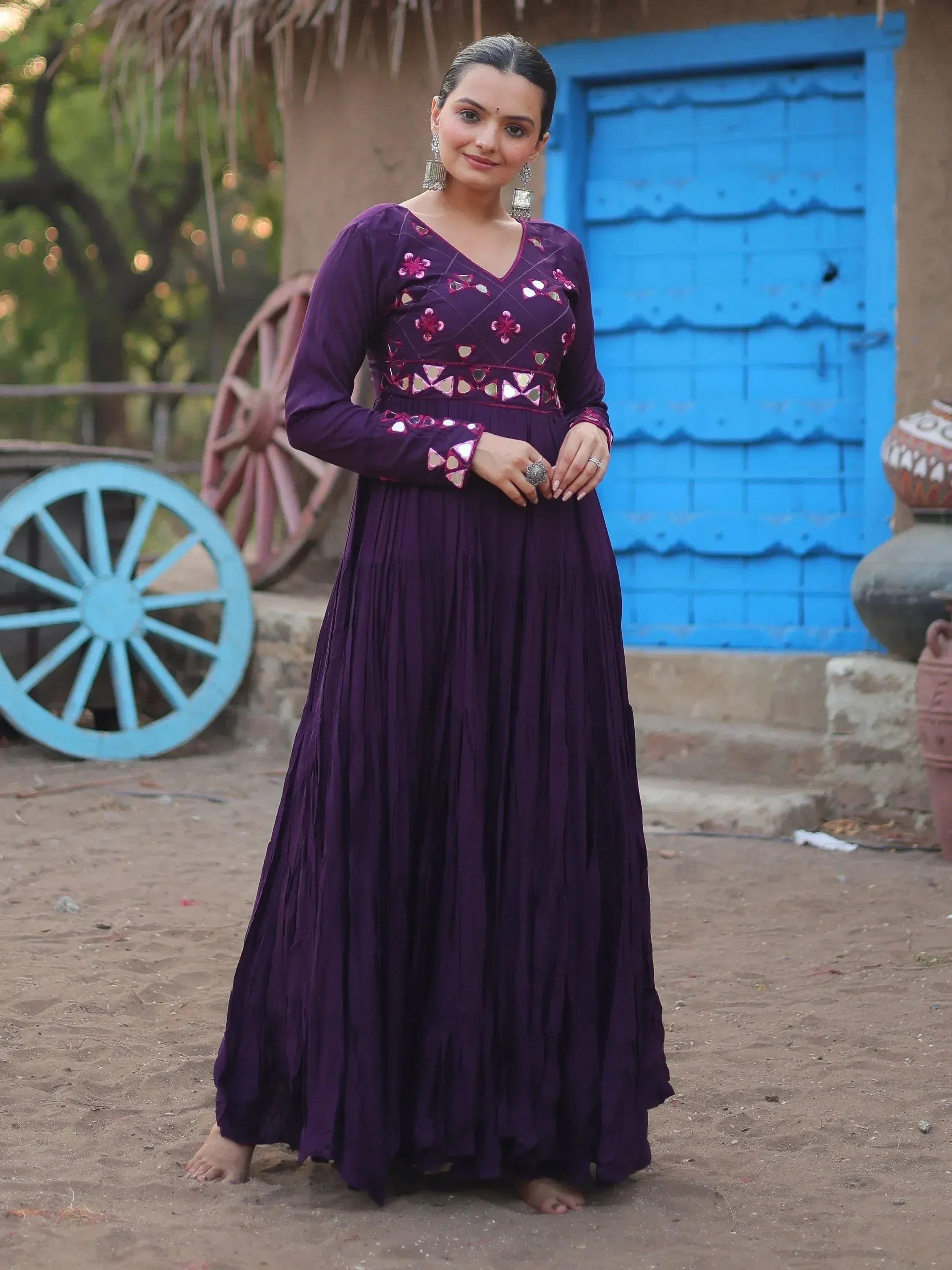 Purple ethnic gown with mirror work, long sleeves, and pleated skirt, styled with traditional jewelry.