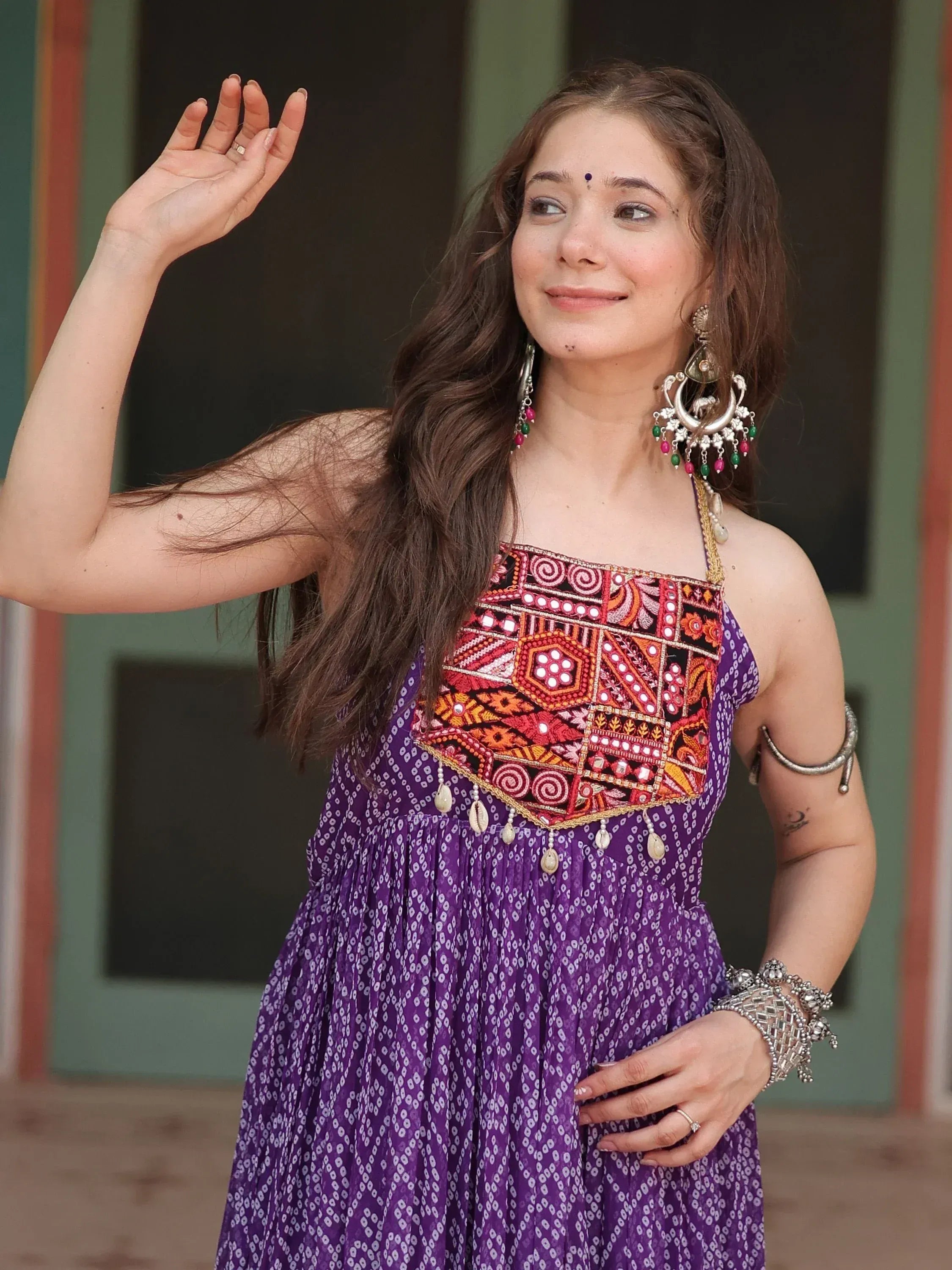Woman in purple ethnic gown with colorful mirror work and traditional jewelry, waving gracefully.