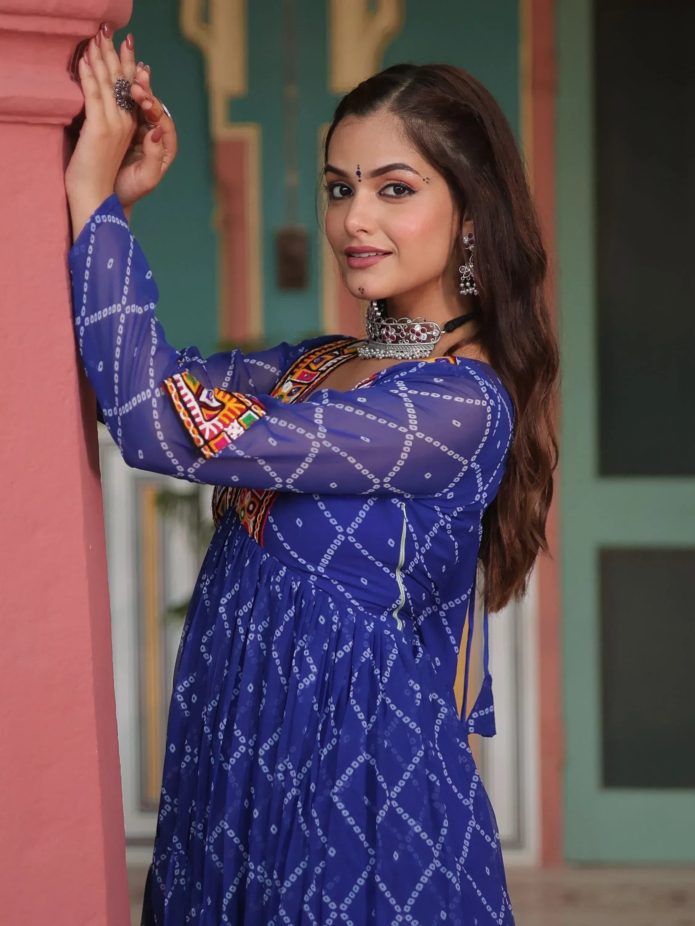 Woman in blue ethnic gown with mirror work, traditional jewelry, and bandhani print, leaning on pink pillar