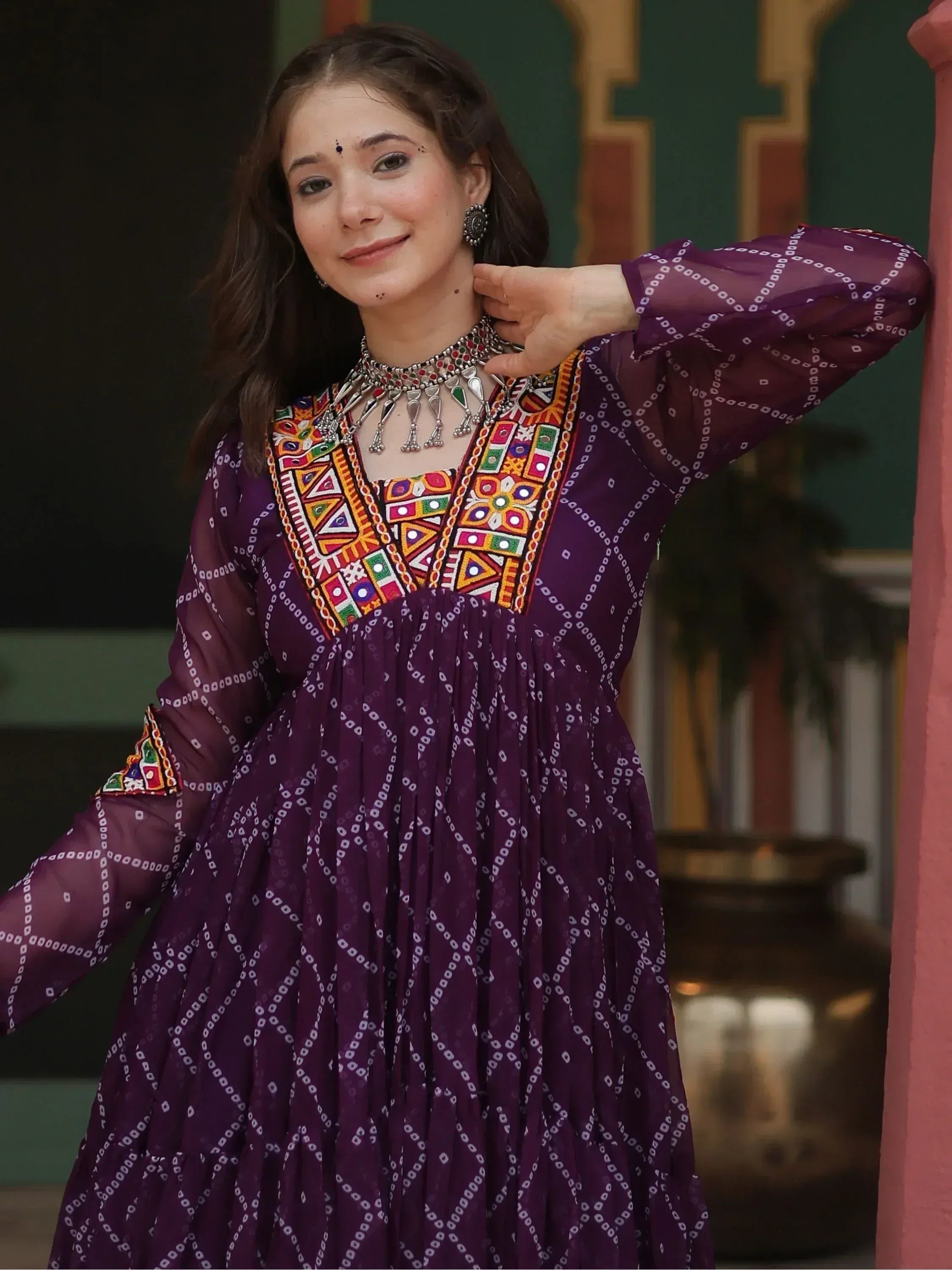 Woman in purple ethnic gown with colorful embroidery and mirror work, styled with traditional jewelry.