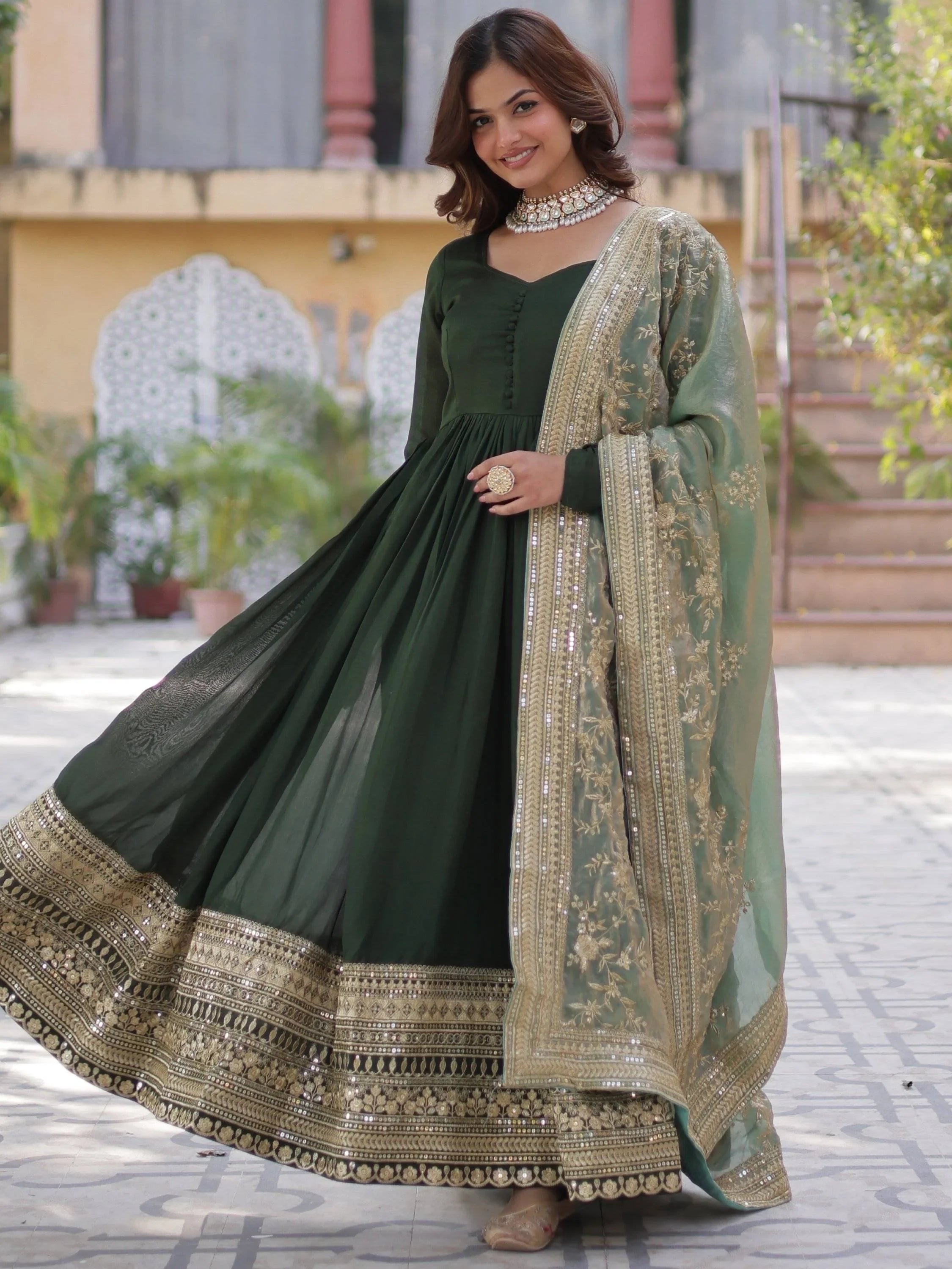 Woman wearing green embroidered ethnic gown with gold dupatta, floral jewelry outdoors