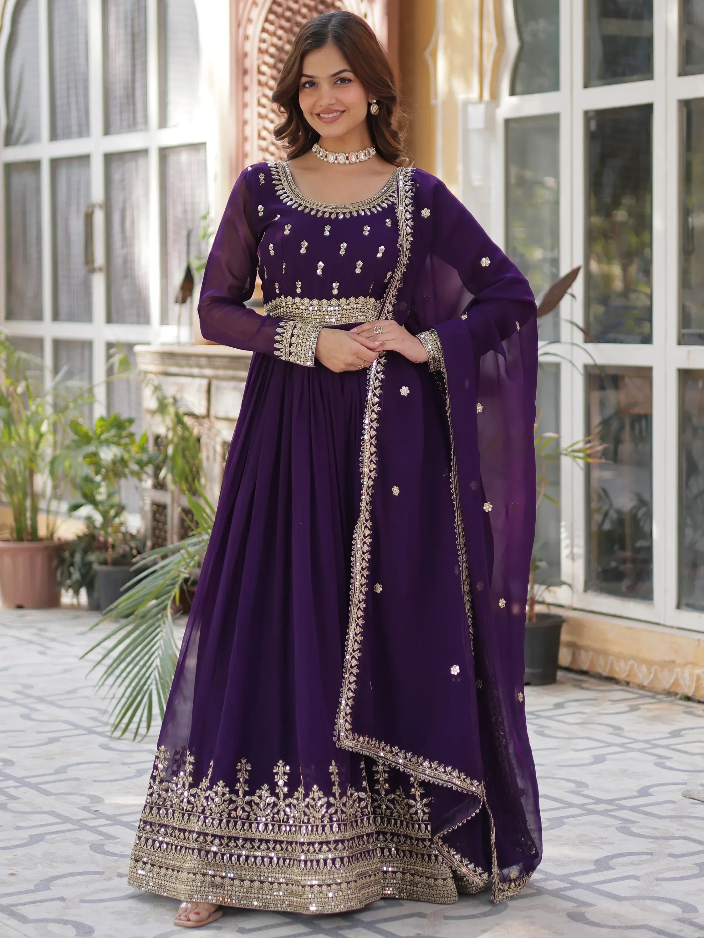 Woman wearing a purple ethnic gown with gold embroidery and dupatta, standing outdoors.