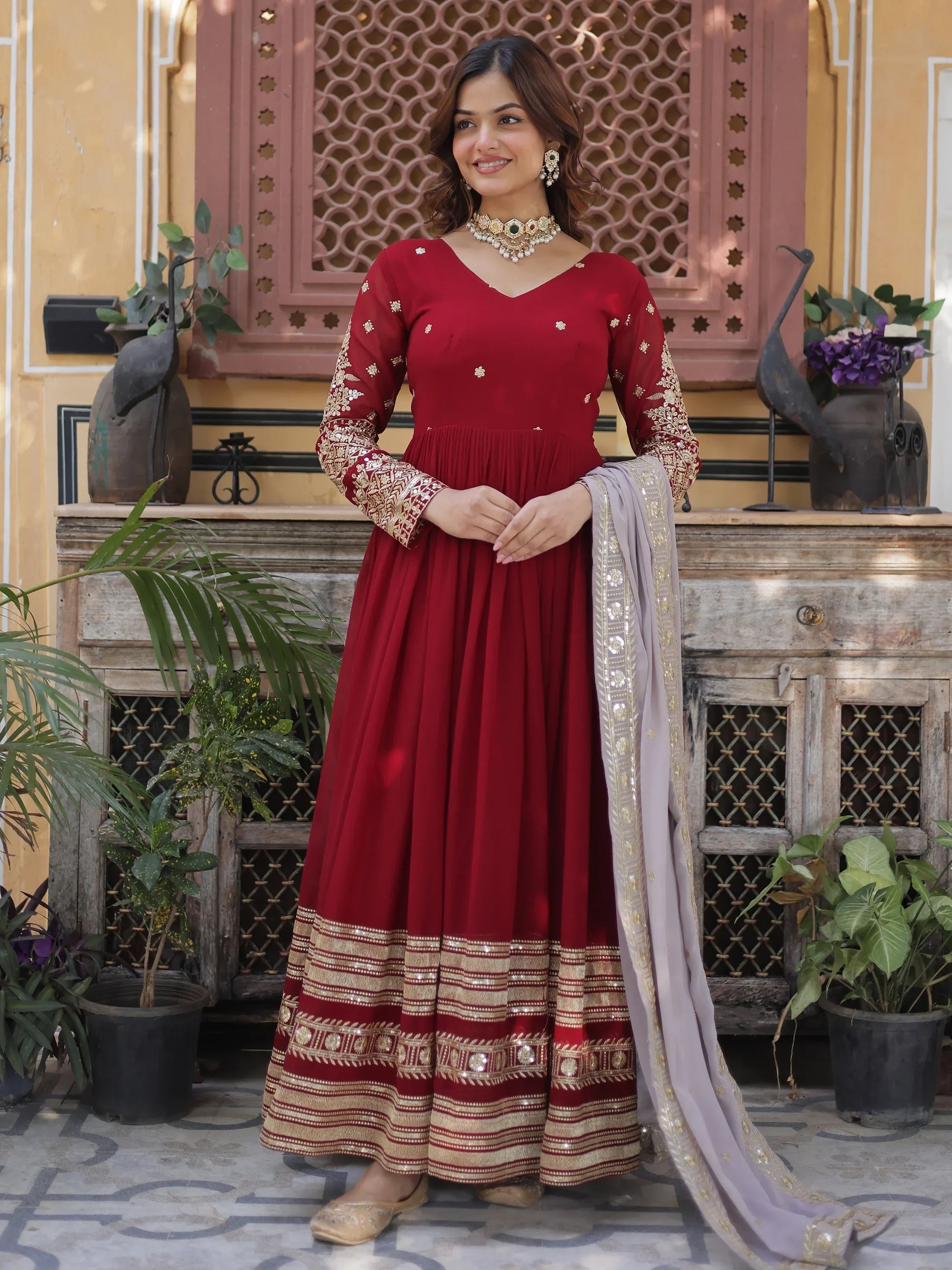 Woman in maroon embroidered Anarkali gown with gold detailing and dupatta, standing outdoors