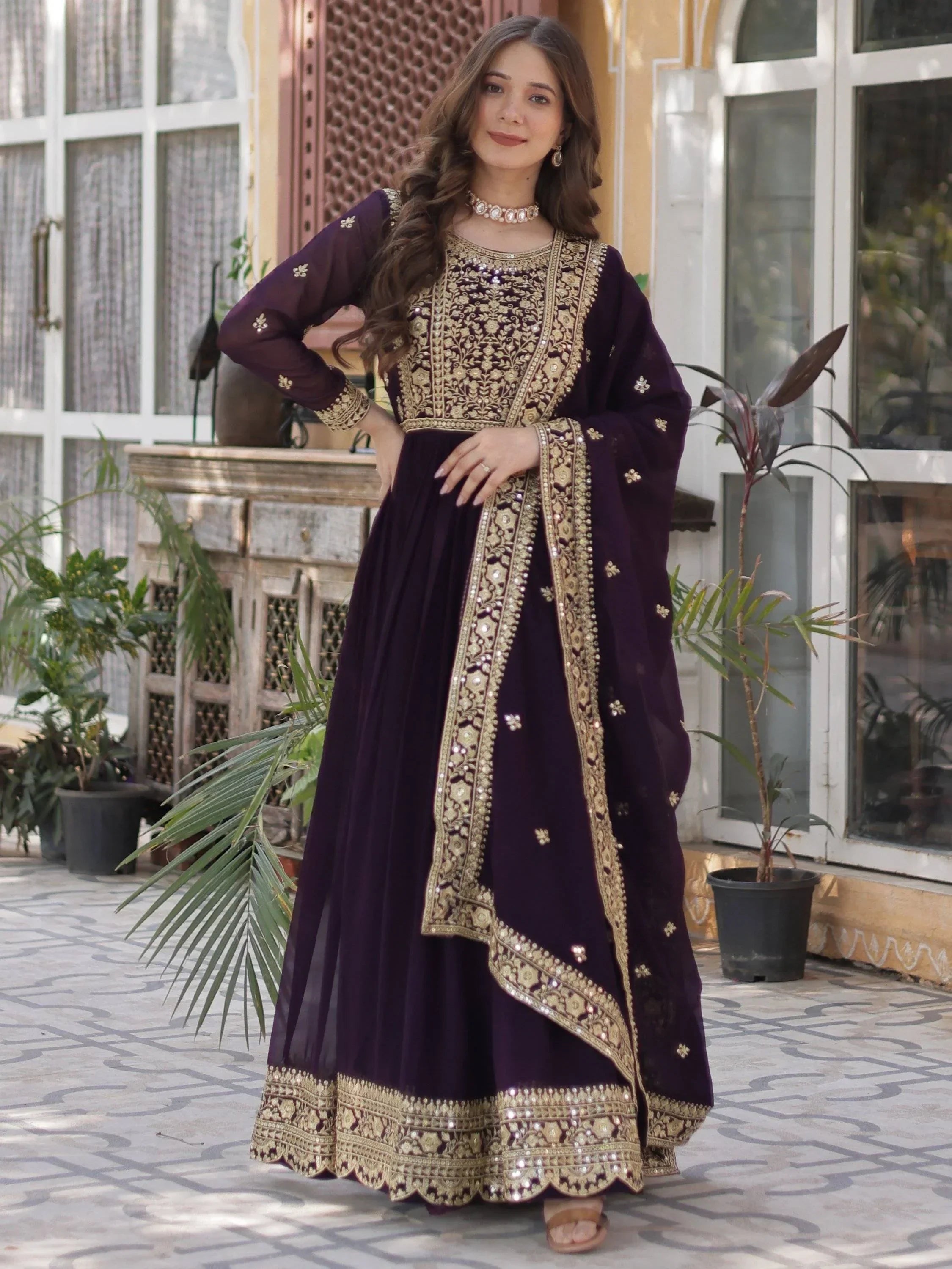 Woman wearing a wine-colored embroidered Anarkali gown with gold detailing, standing outdoors