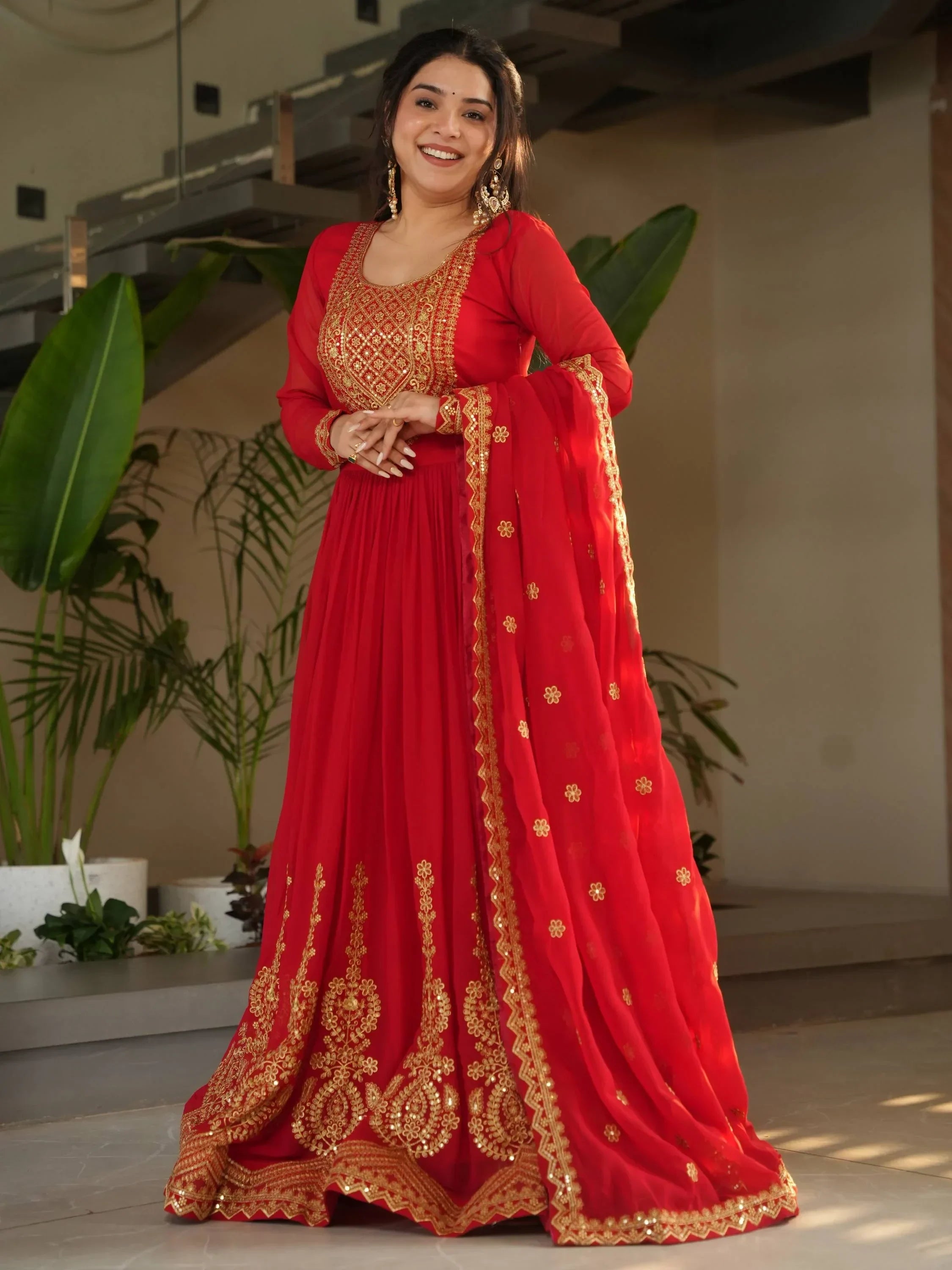 Woman wearing red and gold embroidered ethnic gown, long sleeves, dupatta, standing indoors