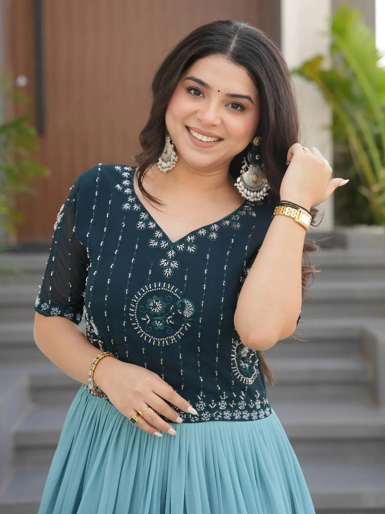 Woman in teal embroidered ethnic gown with pleated skirt, statement earrings, and gold jewelry