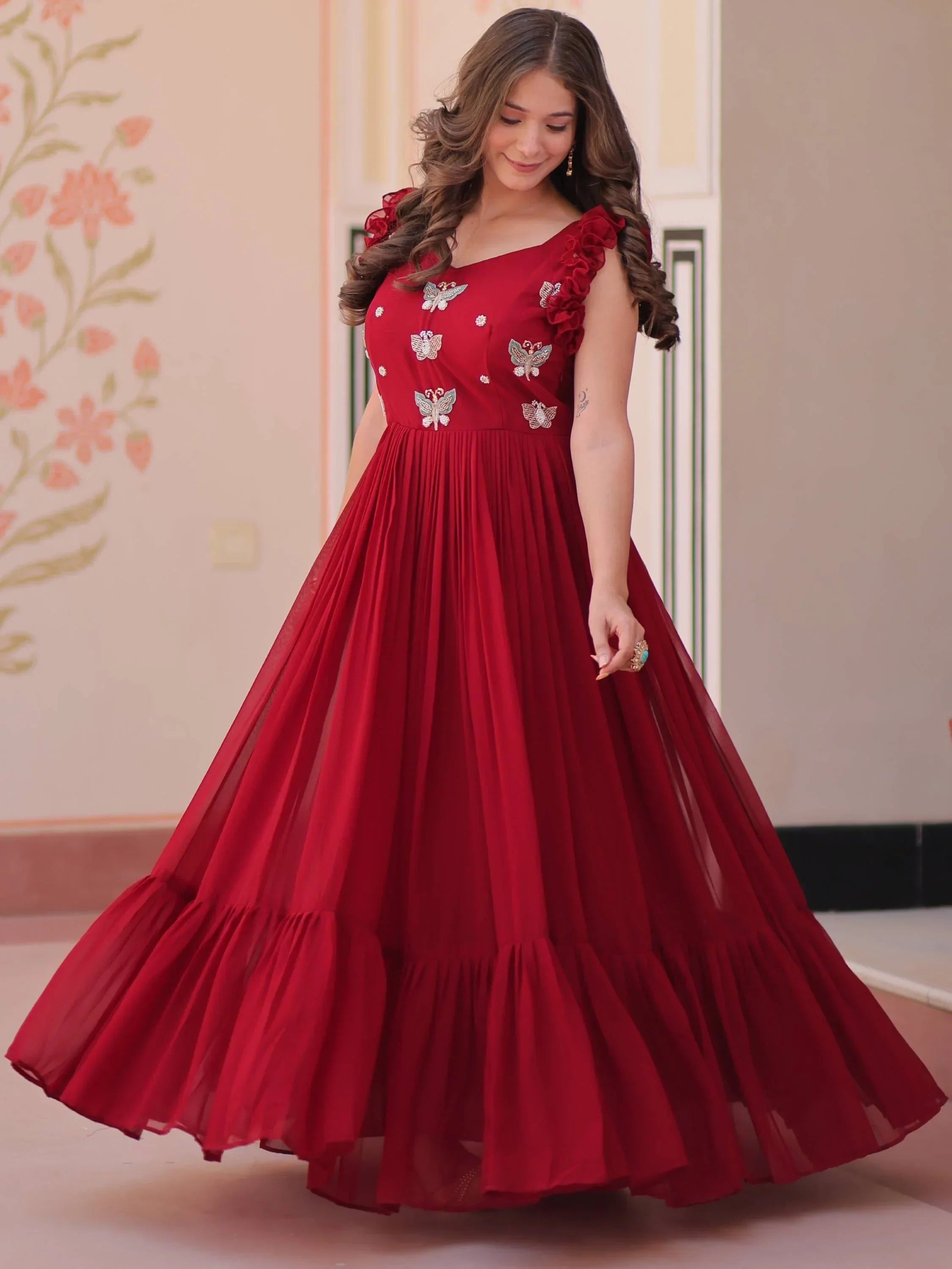 Woman in maroon ethnic gown with butterfly embroidery and ruffle sleeves, standing indoors