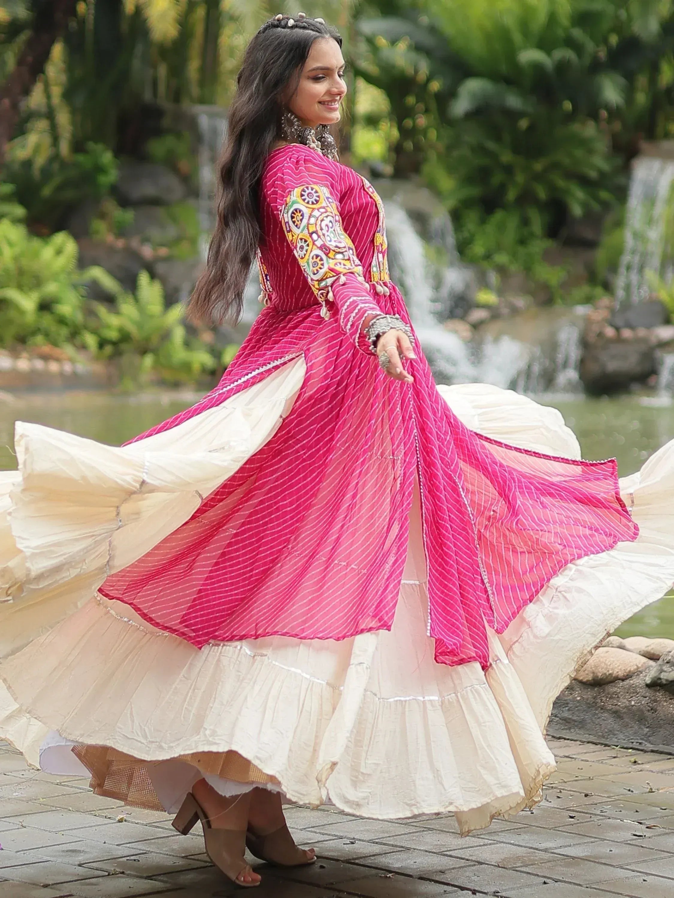 Woman twirling in vibrant pink and cream ethnic gown with embroidered sleeves, set against a lush garden backdrop.