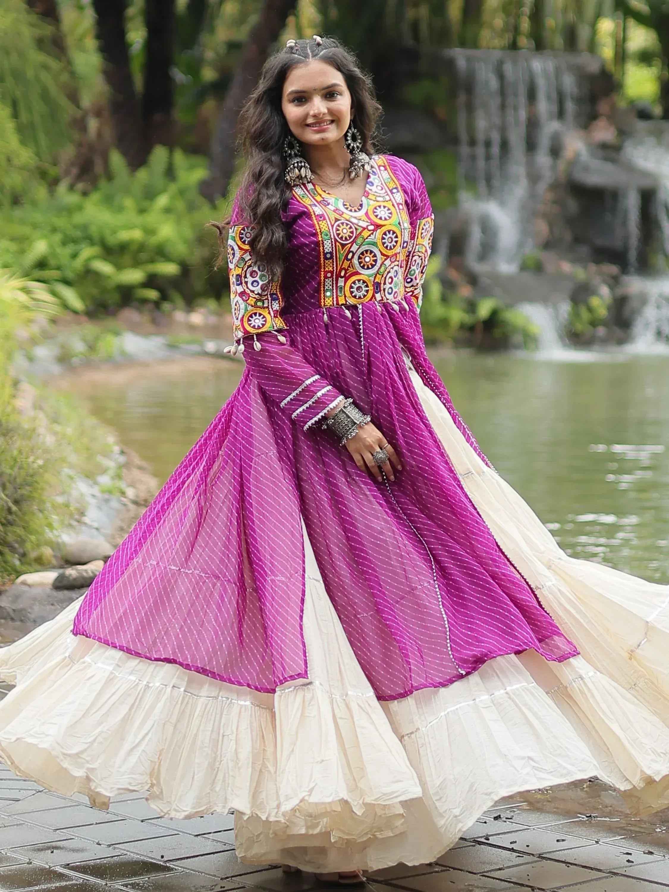 Woman in a purple and cream ethnic gown with mirror work, outdoors by a waterfall