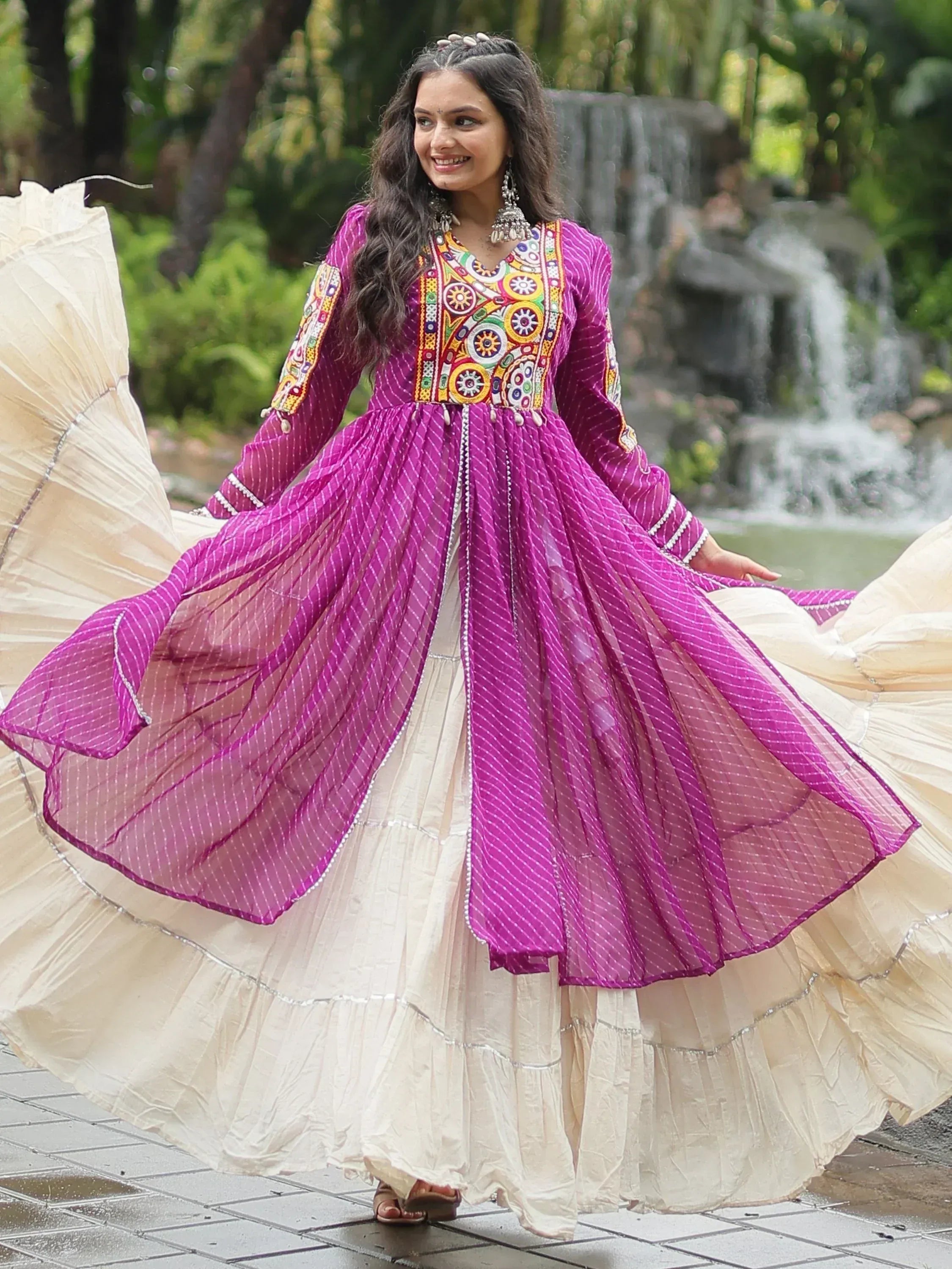 Woman in embroidered magenta ethnic gown with cream lehenga skirt, outdoor waterfall background