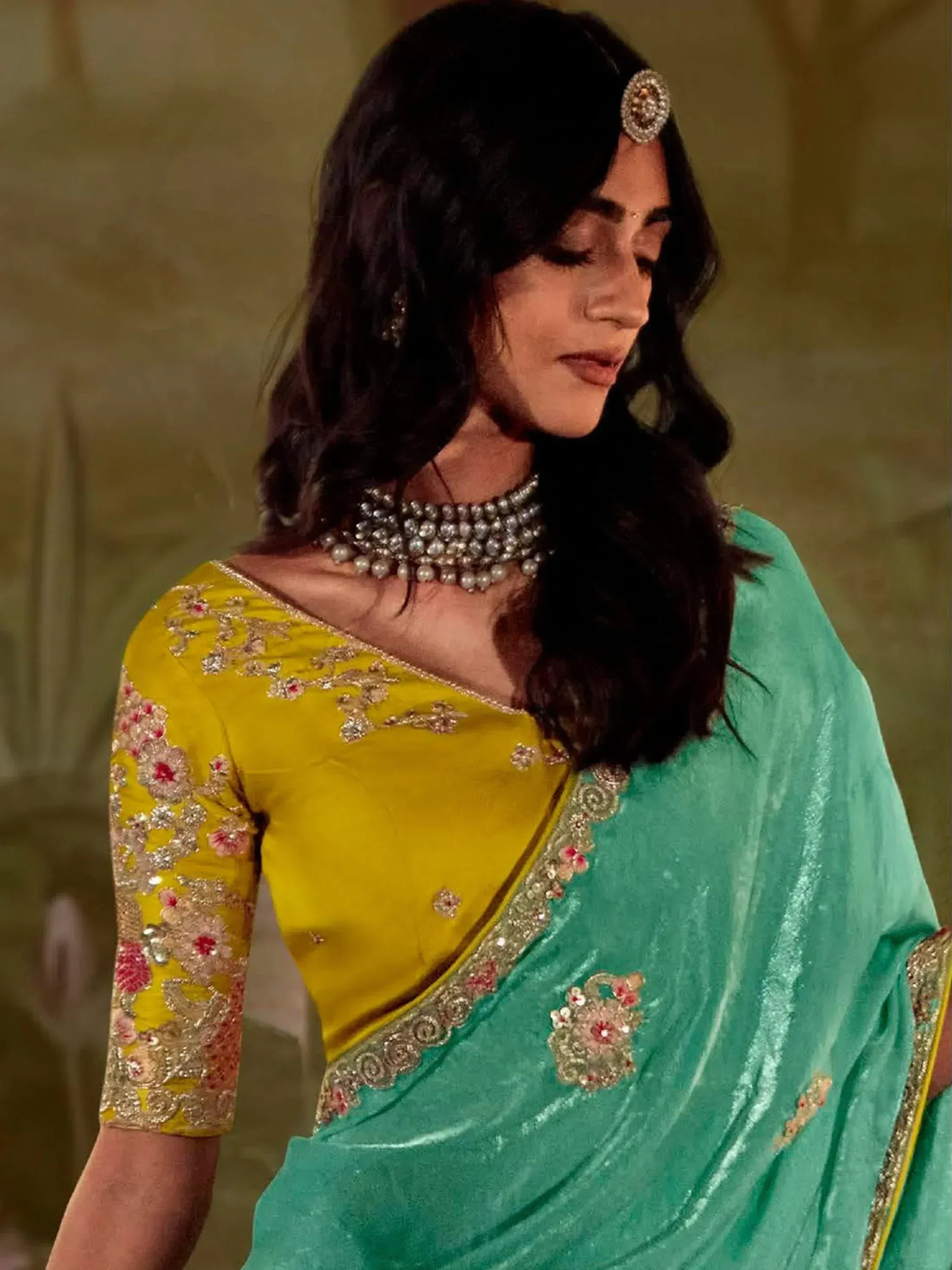 Woman in turquoise velvet saree with yellow embroidered blouse, floral embellishments, and pearl jewelry