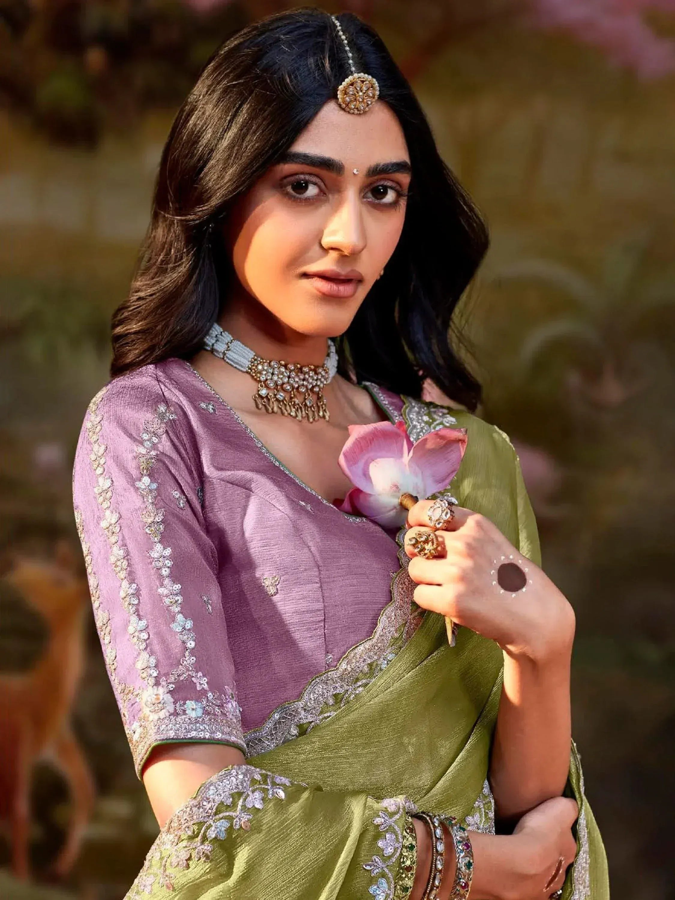 Woman in purple and green embroidered saree with traditional jewelry, holding pink flower