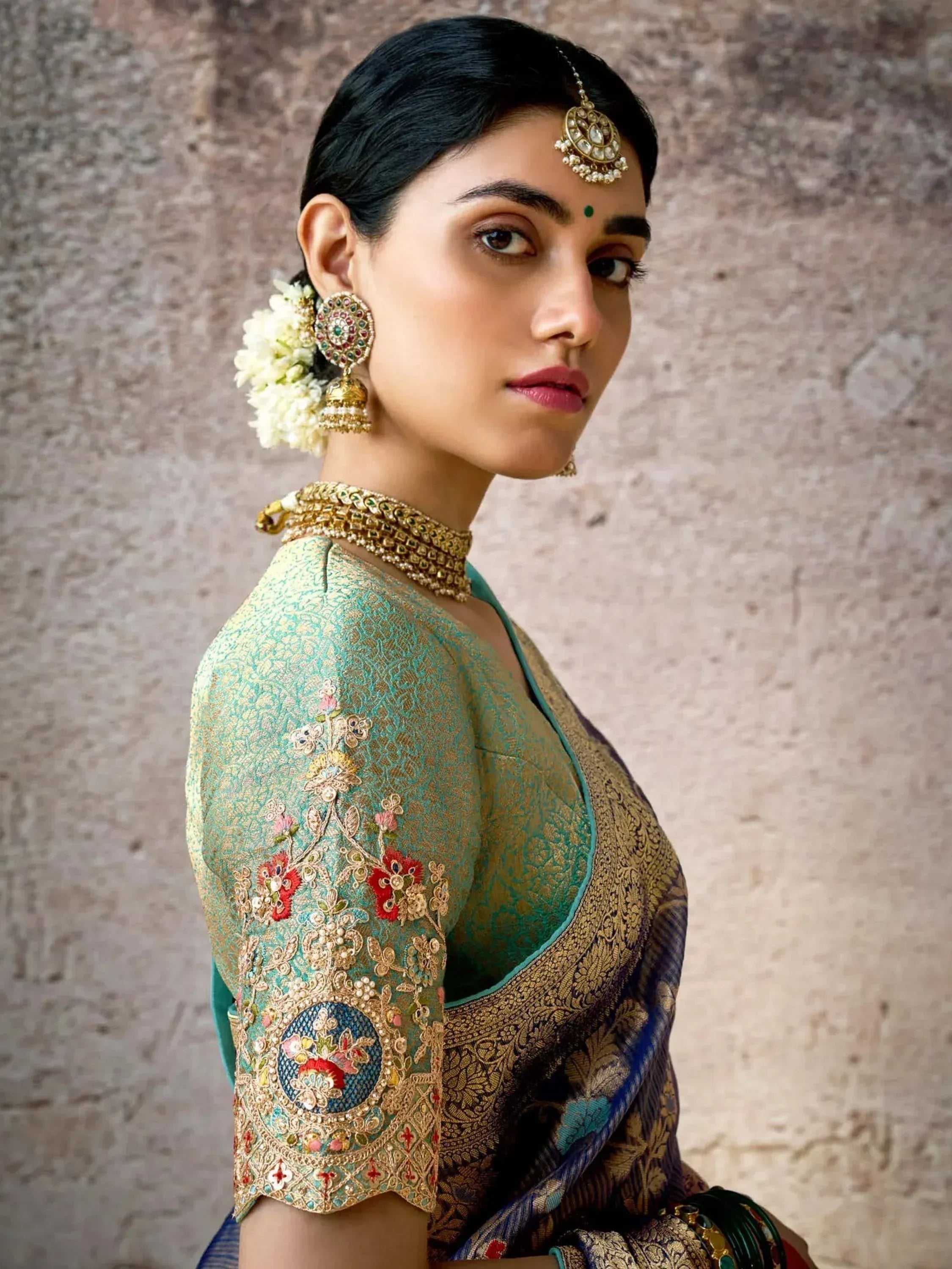 Elegant woman in teal embroidered saree blouse with gold jewelry and floral hair accessory
