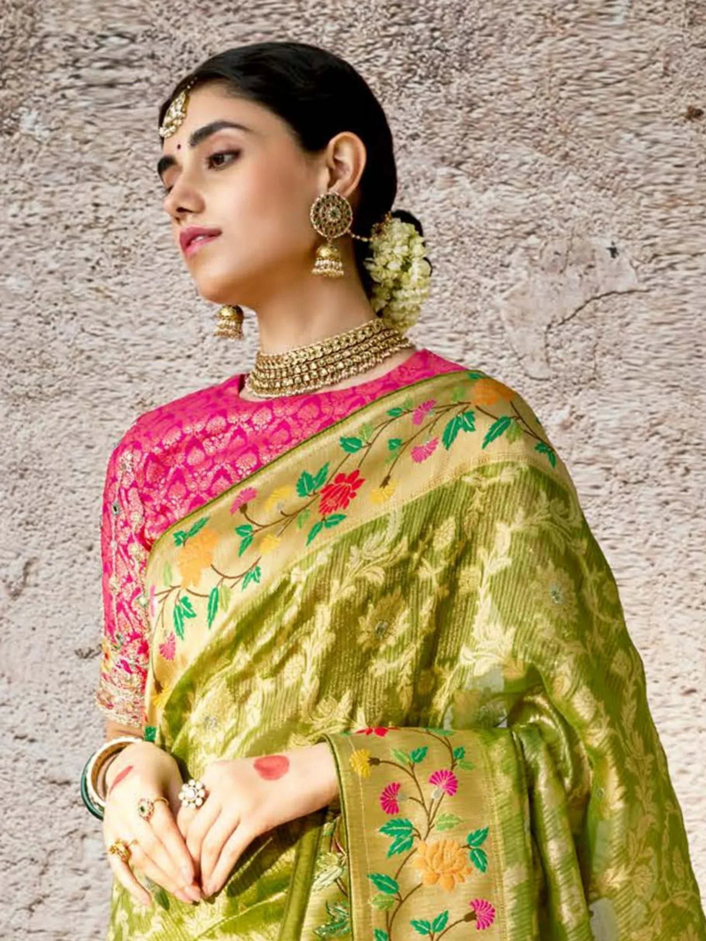 Woman in vibrant green floral saree with pink blouse, traditional Indian ethnic wear, gold jewelry, and floral hair accessory.