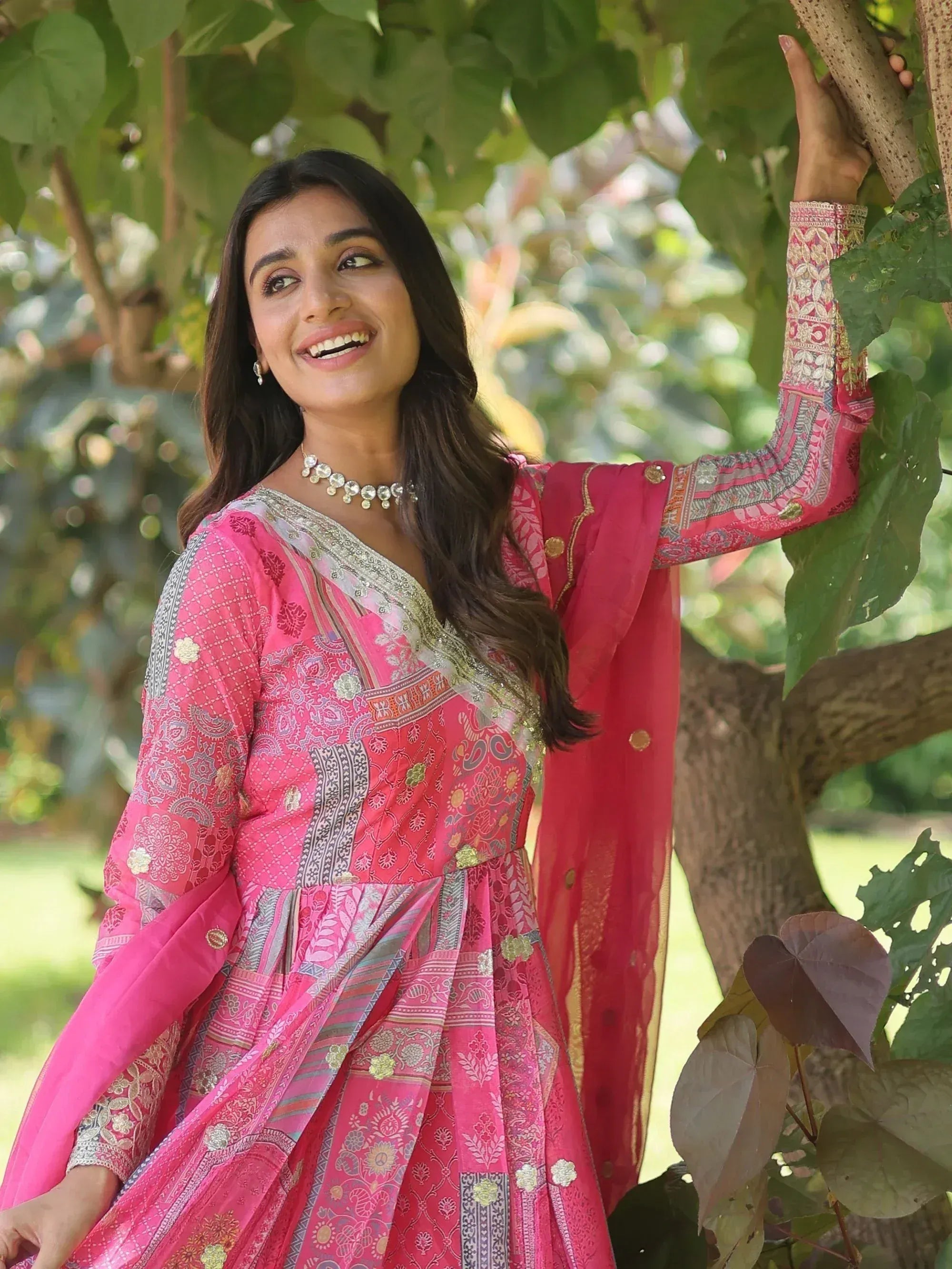 Woman in pink printed ethnic gown with gold embroidery, standing under tree, perfect for festive occasions