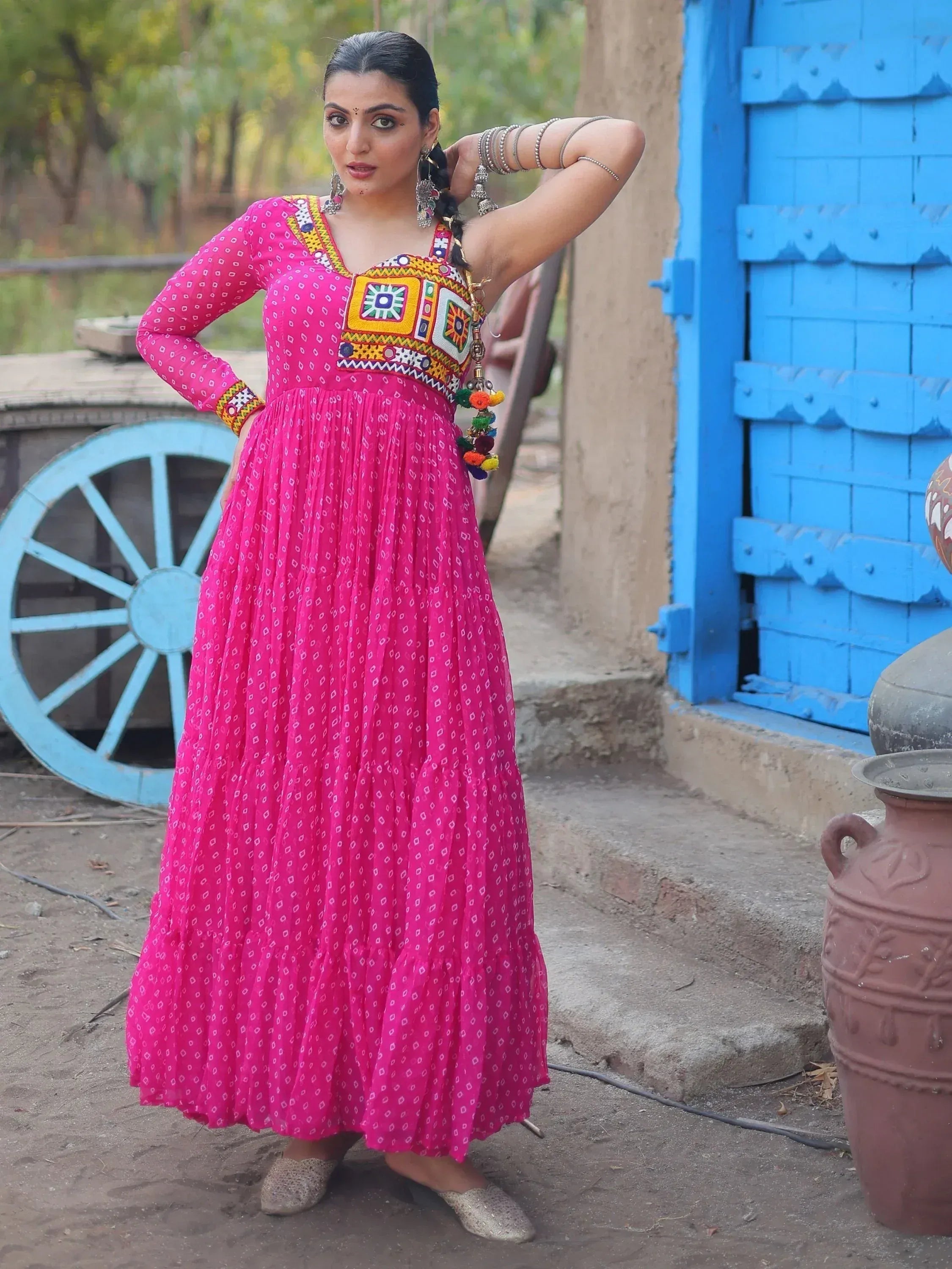 Woman in vibrant pink ethnic gown with mirror work, standing by blue door and cart wheel