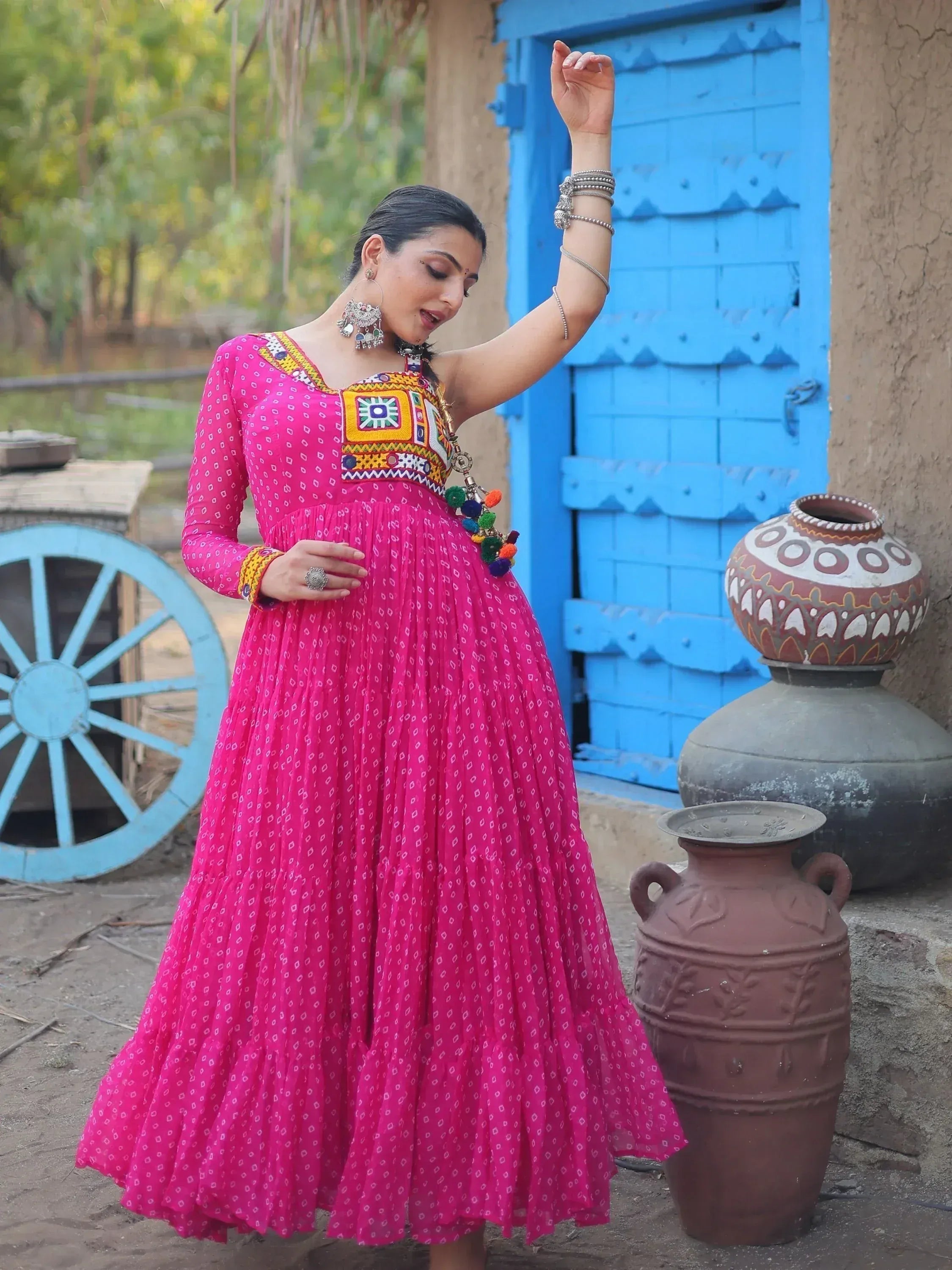 Woman in vibrant pink ethnic gown with mirror work, standing by blue door and clay pots